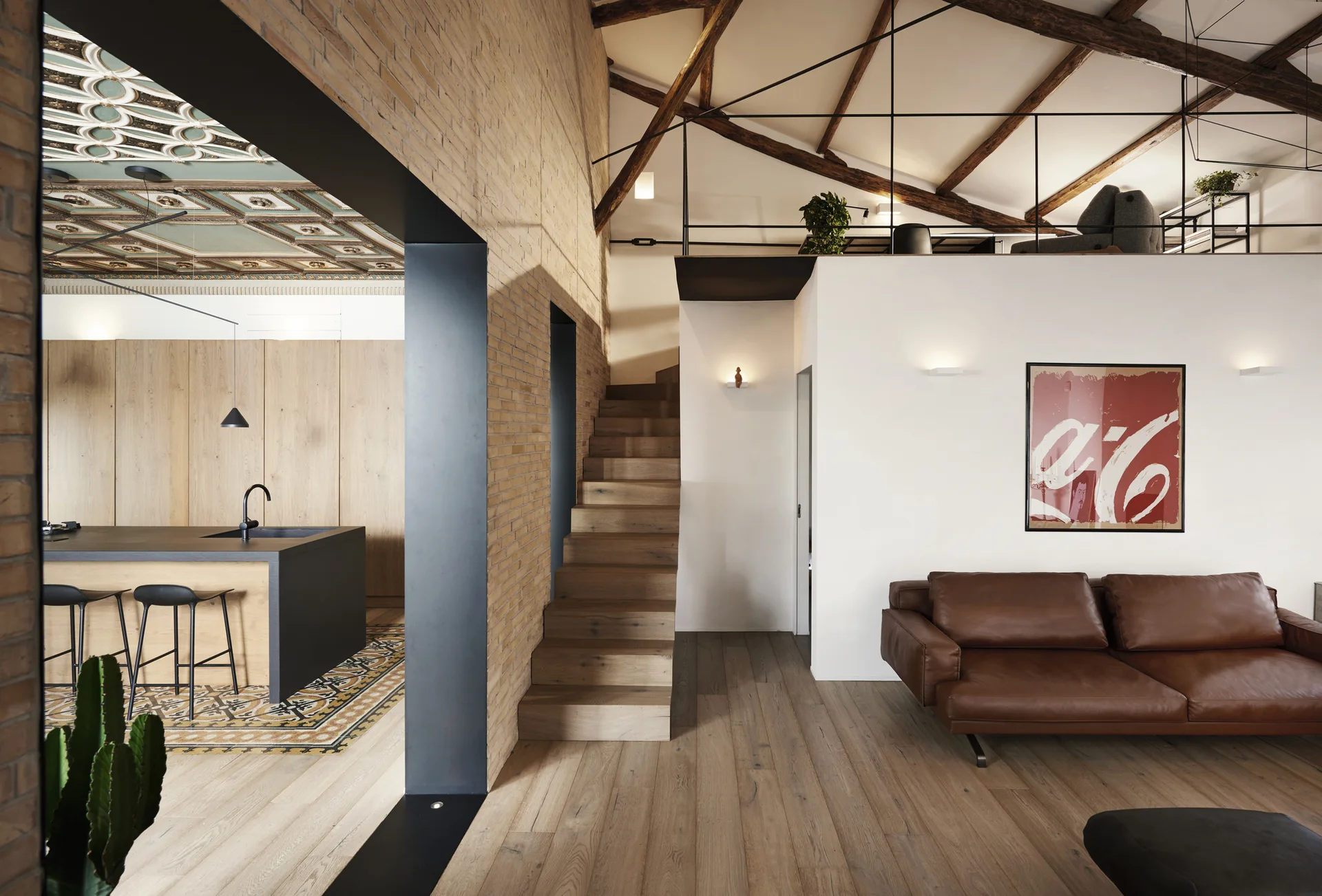 An open-plan loft featuring a brown leather sofa, wooden staircase, exposed brick wall, and a kitchen with wood cabinets.