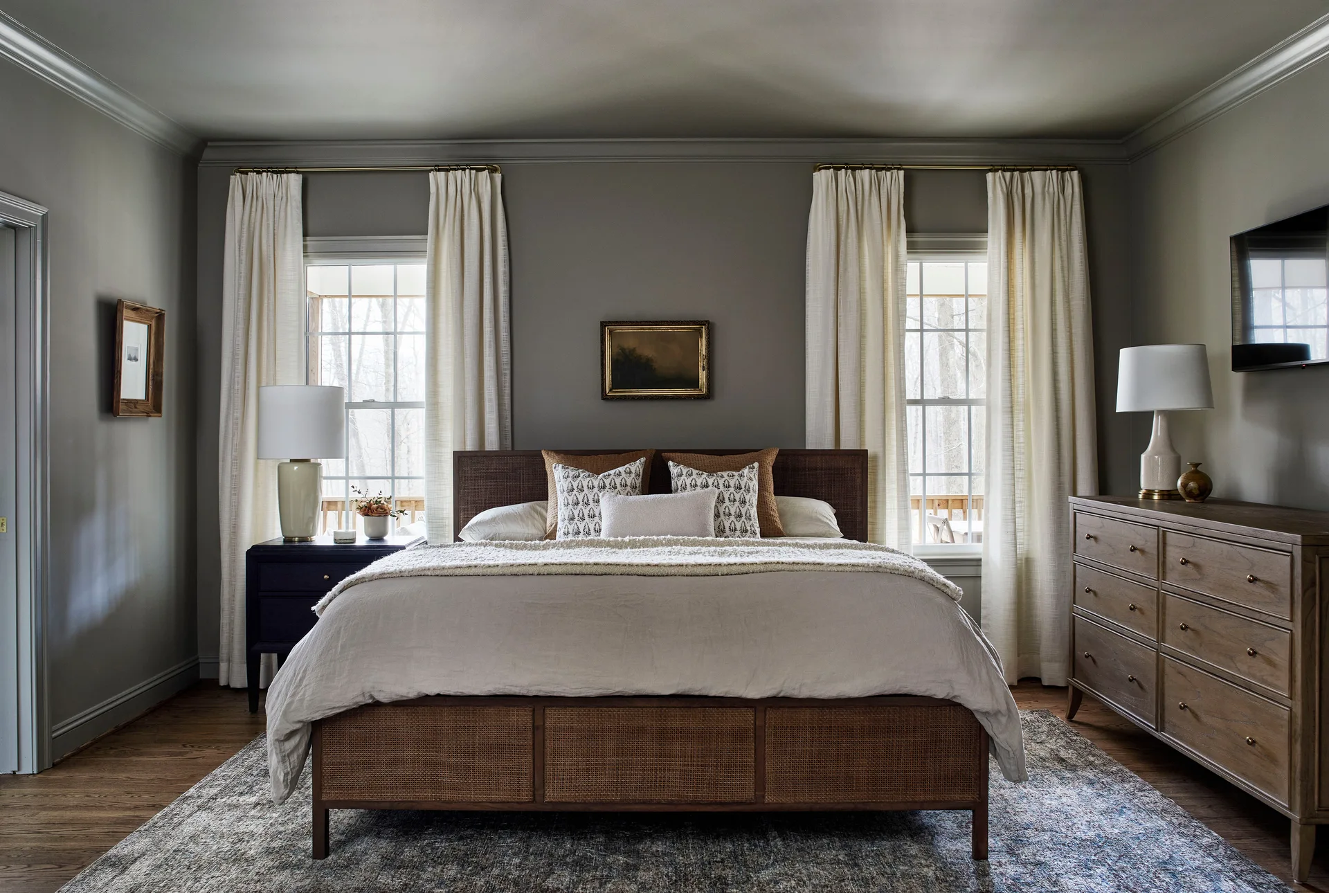 A bedroom featuring a wooden bed with a cane headboard set between two windows with white drapes against sage green walls.