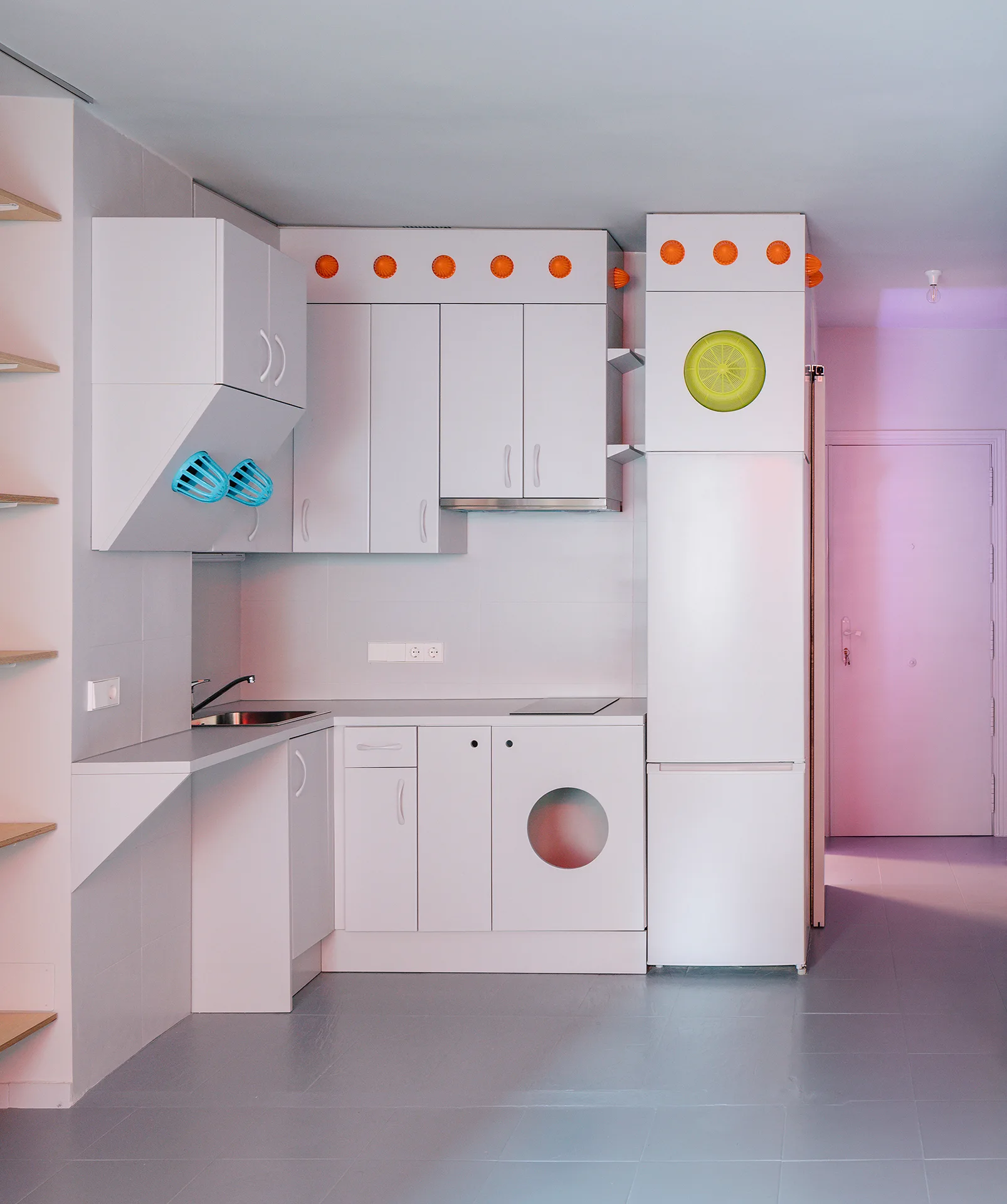 A minimalist white kitchen features matte cabinetry, an integrated washing machine, grey tiled floors, and colorful orange and blue accents.