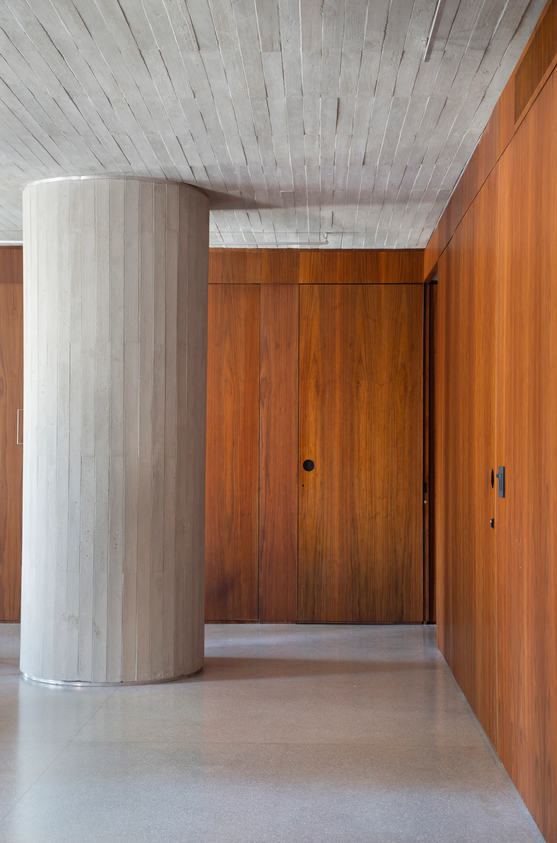A minimalist hallway features a large textured concrete column alongside warm wood-paneled walls with flush doors and a polished floor.