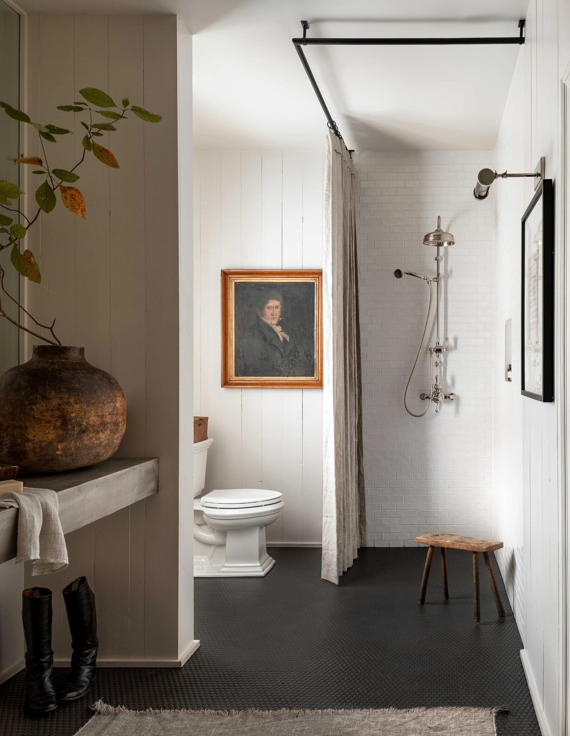 A neutral bathroom features white vertical wood paneling, a subway-tiled shower with a linen curtain, a toilet, and a framed portrait painting.