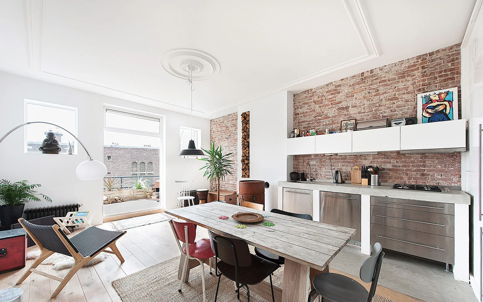 An open-concept kitchen features an exposed brick wall, stainless steel cabinets, and a rustic wooden dining table with mismatched chairs.