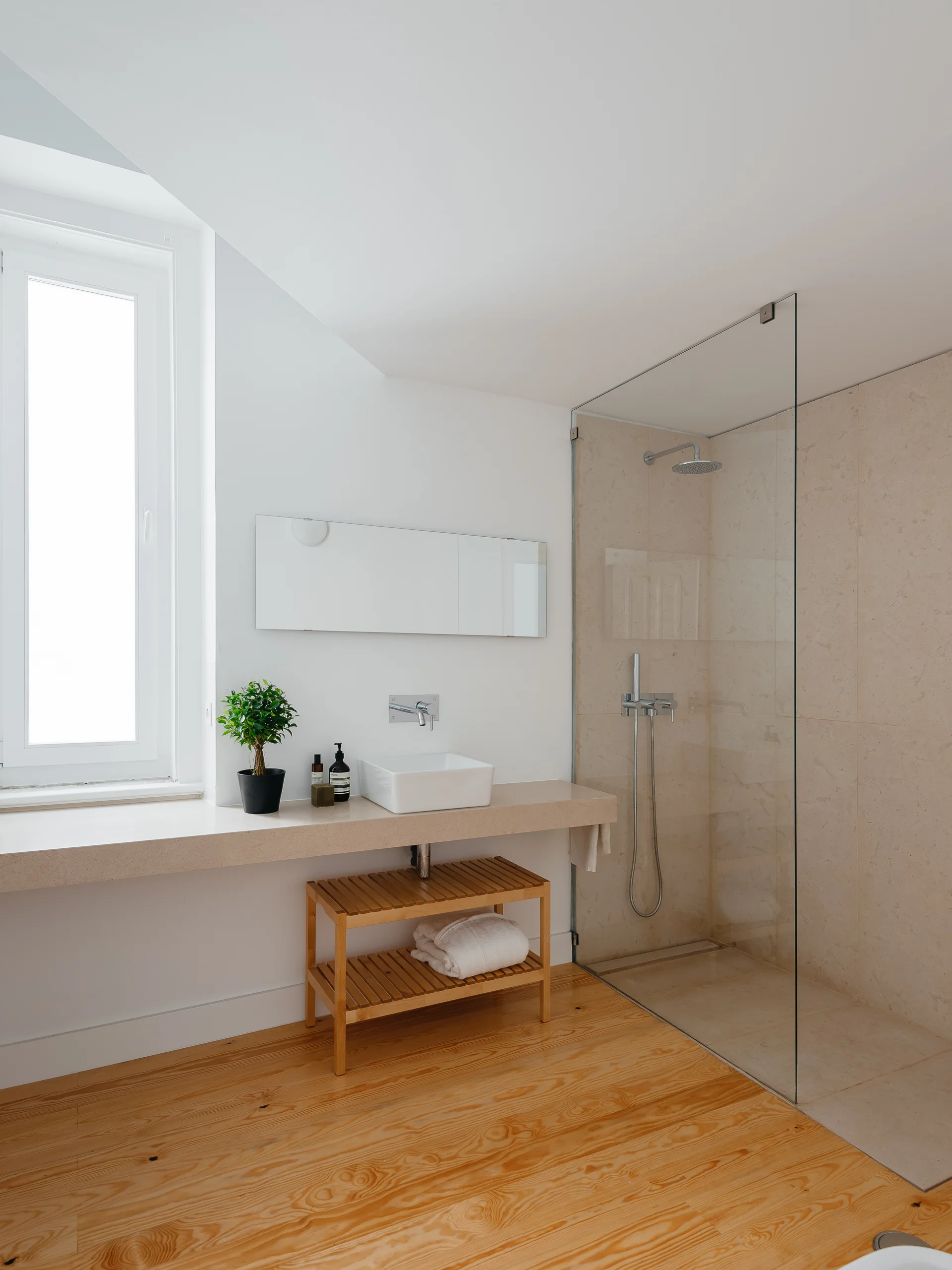 A minimalist bathroom with a long stone vanity ledge, a white vessel sink, a slatted wooden bench underneath, and a glass-enclosed shower.