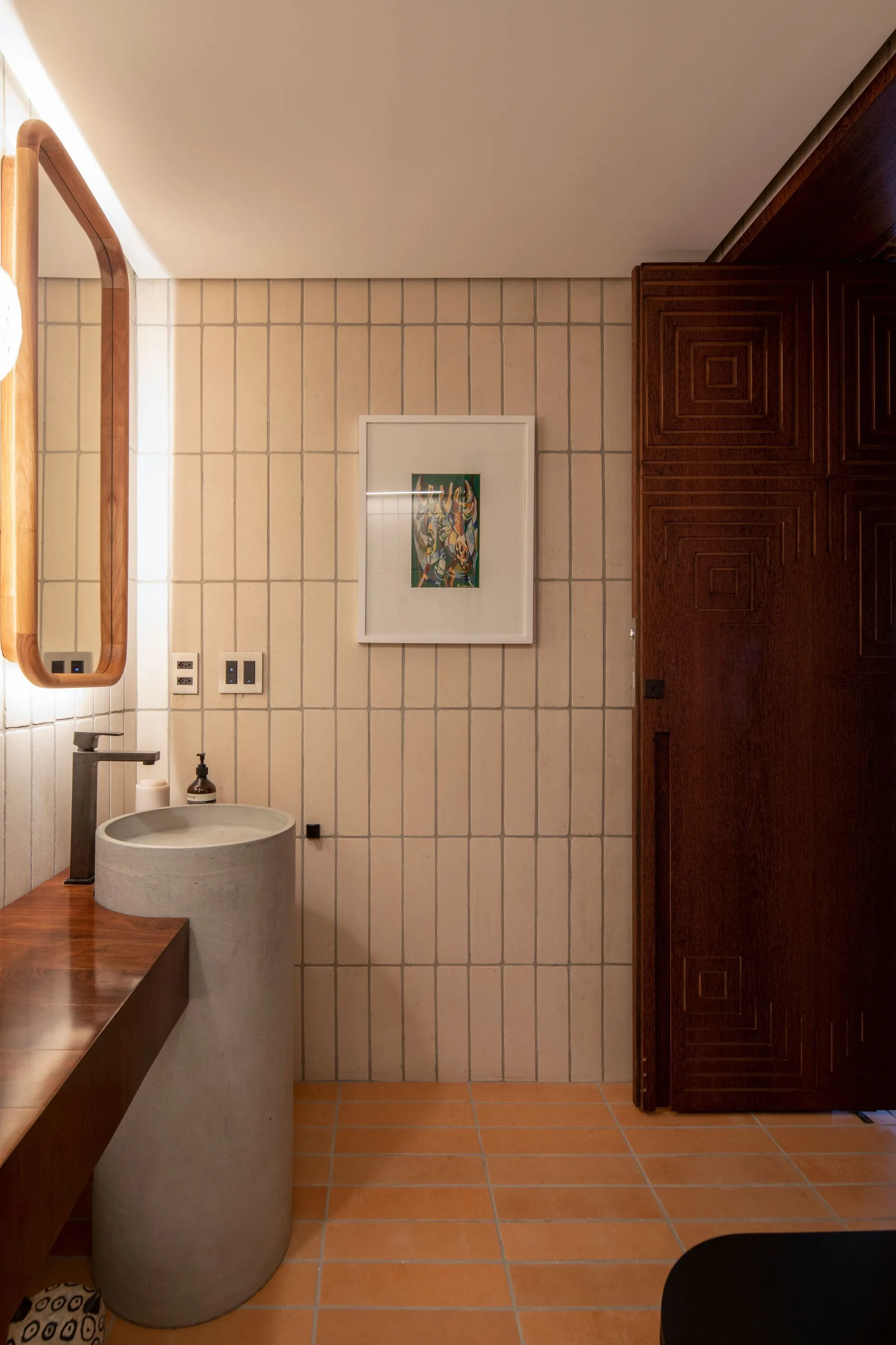 A modern bathroom features a cylindrical concrete pedestal sink, vertical beige tiled walls, a backlit mirror, and a carved wooden door.