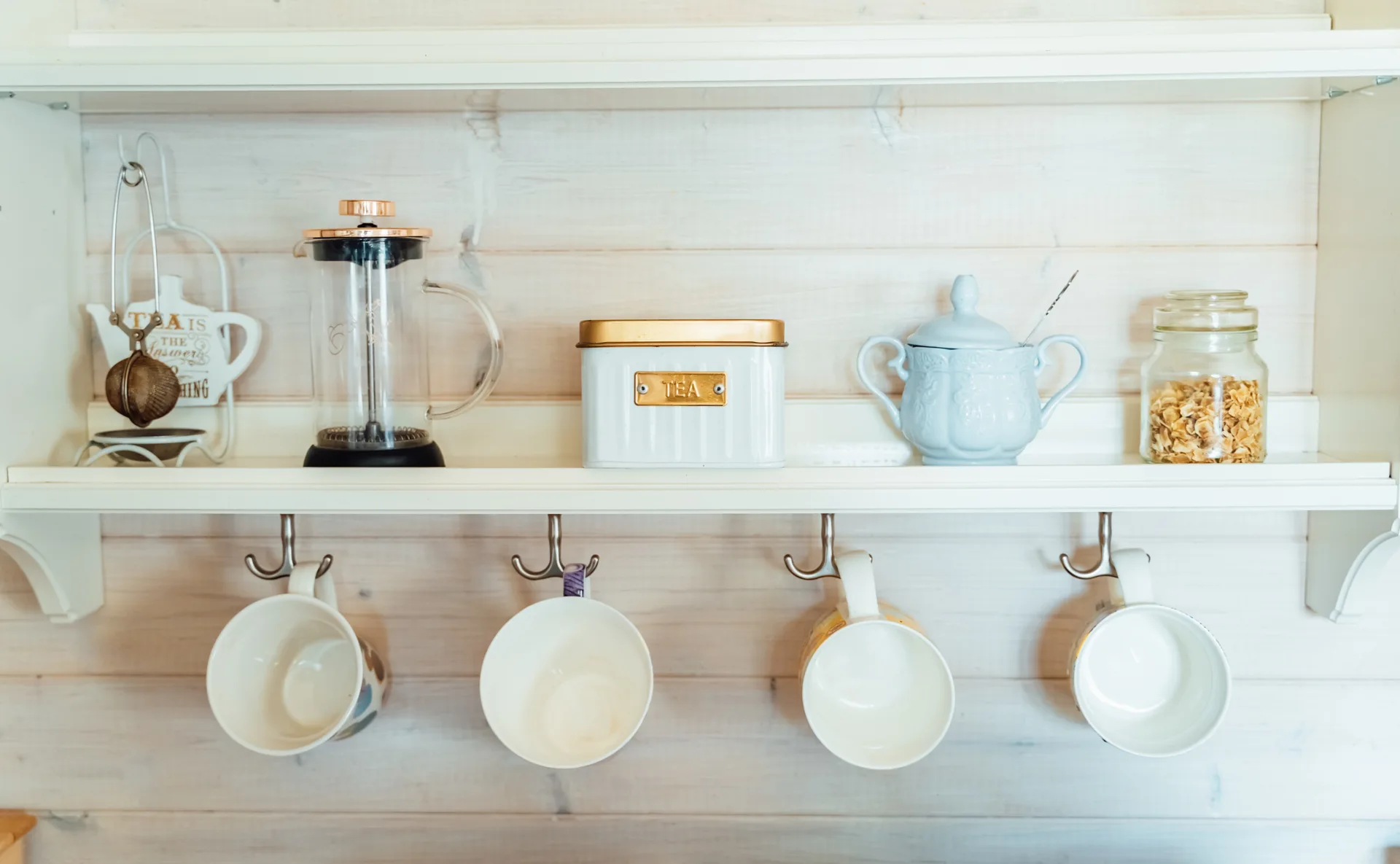 A white kitchen shelf holding a French press, canister, and jar, with four ceramic mugs hanging from metal hooks below against a wood paneled wall.