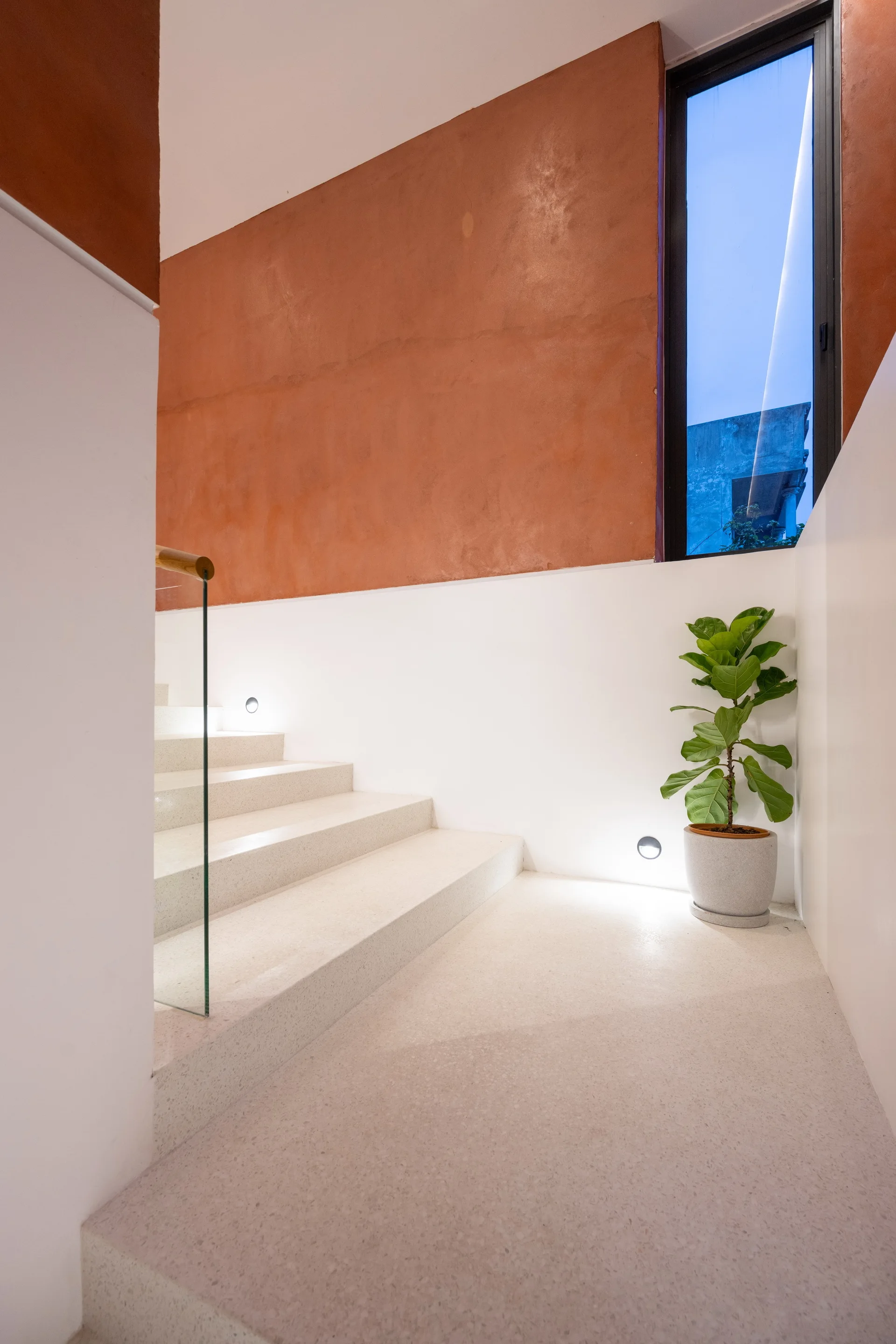 A modern minimalist stairwell features cream terrazzo steps, a glass railing, and a terracotta-colored upper wall accented by a potted plant.