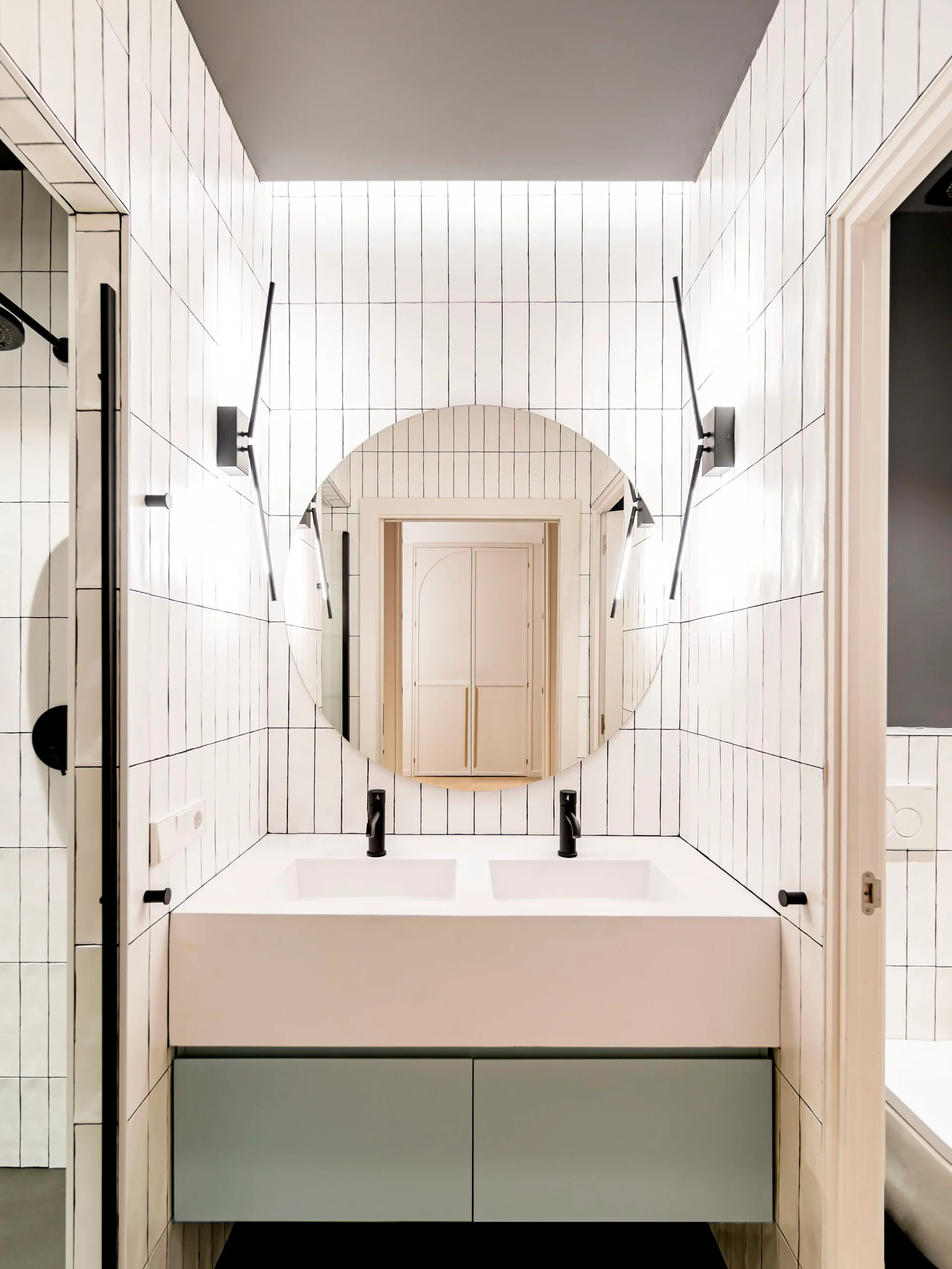 A modern bathroom features a double sink vanity, a large round mirror, black faucets, and walls covered in vertical white tiles with dark grout.