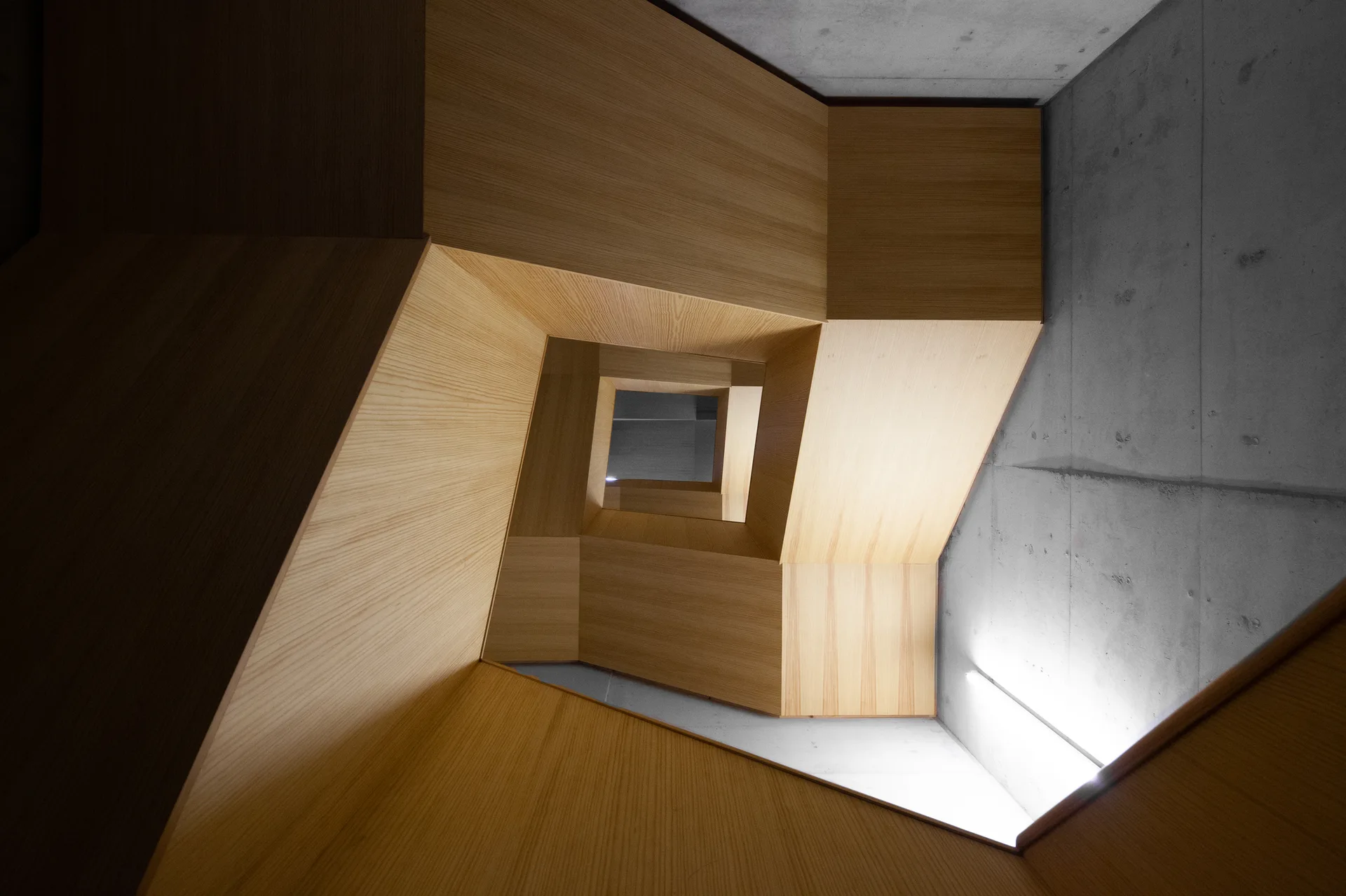 Looking down a square spiraling staircase featuring light wood-toned paneling alongside an exposed grey concrete wall.