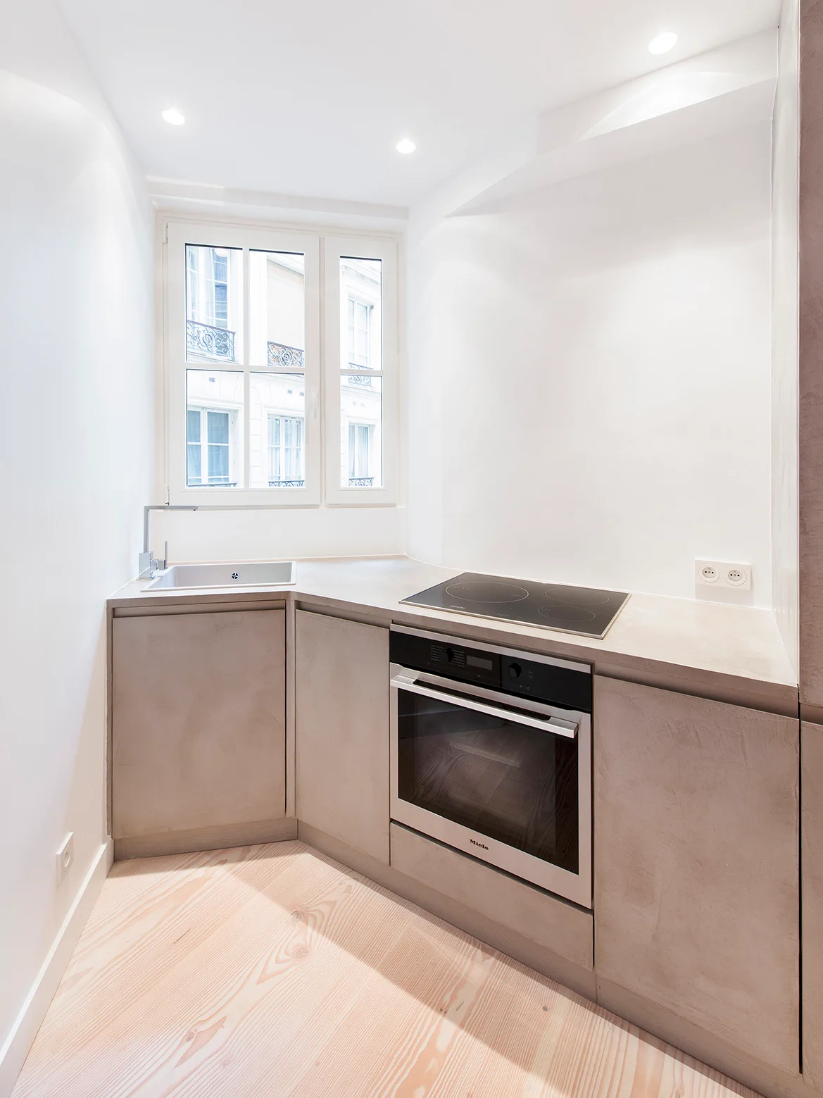 A small minimalist kitchen features matte beige cabinetry, an angled countertop with a sink, an oven, and light wood flooring.