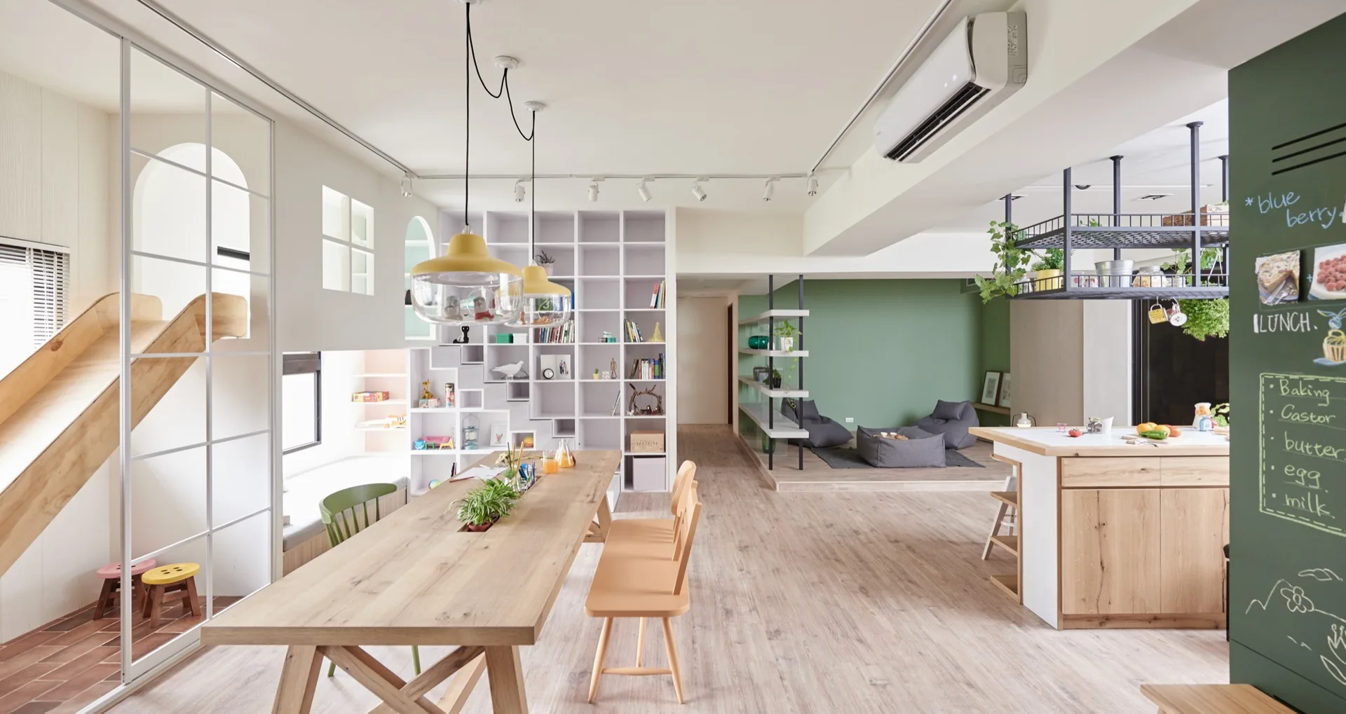 An open-concept living area features a wooden dining table, a large white bookshelf, and a custom indoor slide next to a kitchen island.