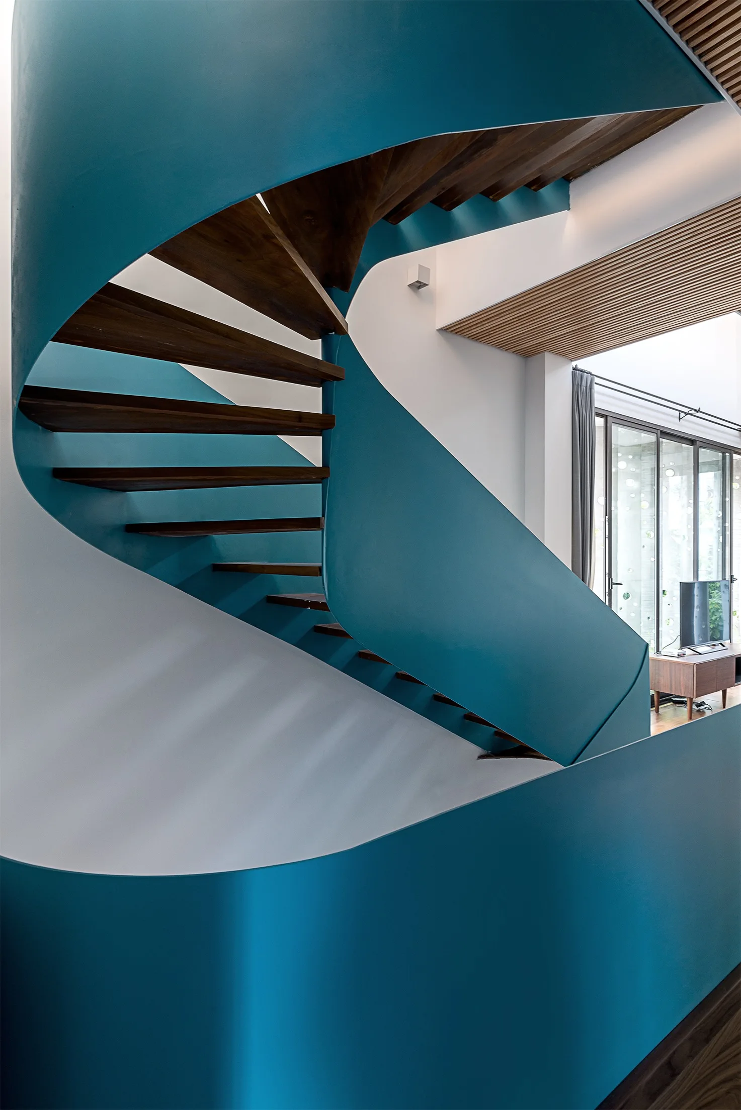 A modern spiral staircase with a curved teal metal outer shell and dark wood treads rises toward a wood-slatted ceiling.
