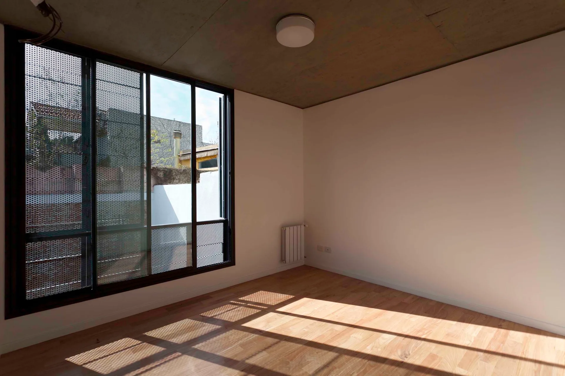 An empty room features light wood flooring, white walls, an exposed concrete ceiling, and a large window covered in perforated metal screens.