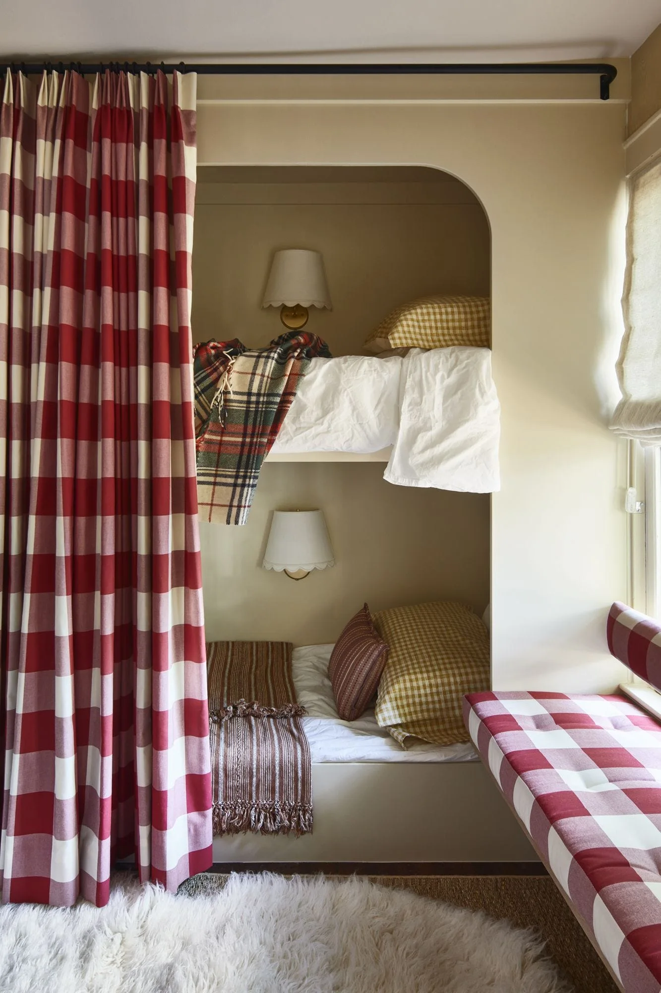 Built-in bunk beds set inside a cream arched alcove with red checkered curtains, scalloped wall sconces, and a matching window seat cushion.