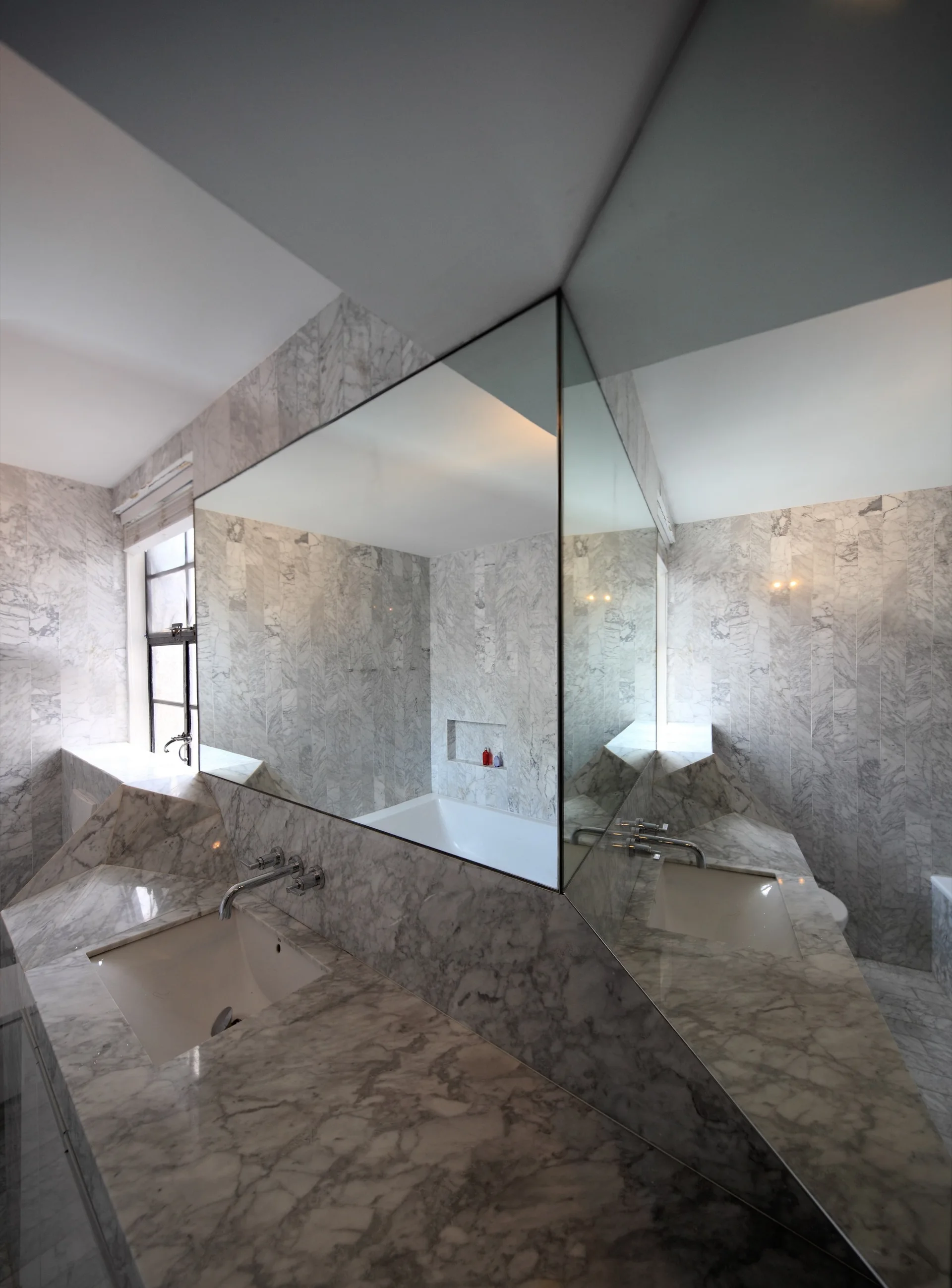 A modern bathroom featuring walls and an angular double vanity clad in grey and white veined marble, topped by a large frameless corner mirror.