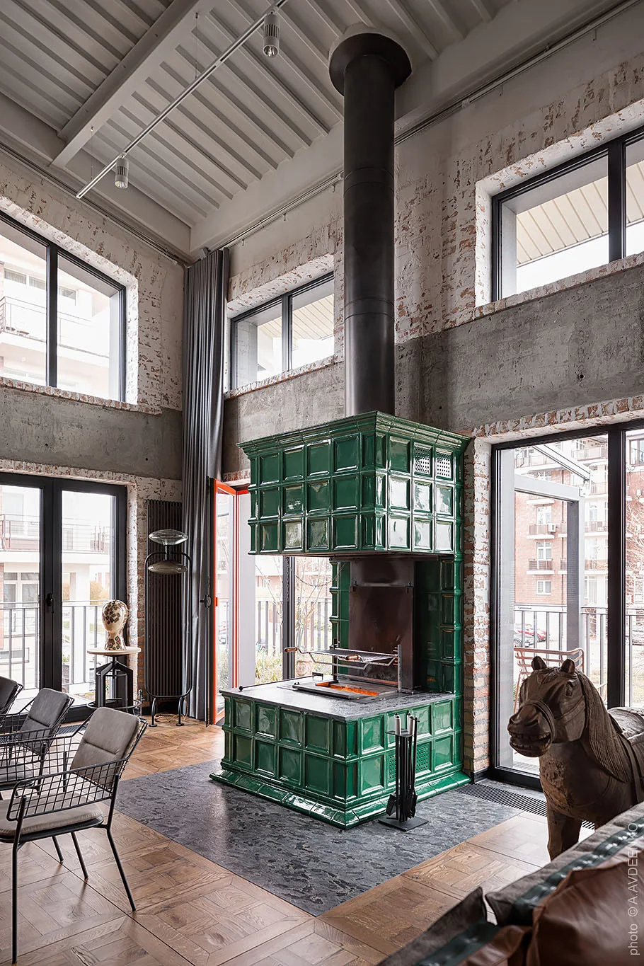A spacious loft living room featuring a glossy green tiled fireplace, exposed brick walls, and tall industrial windows.