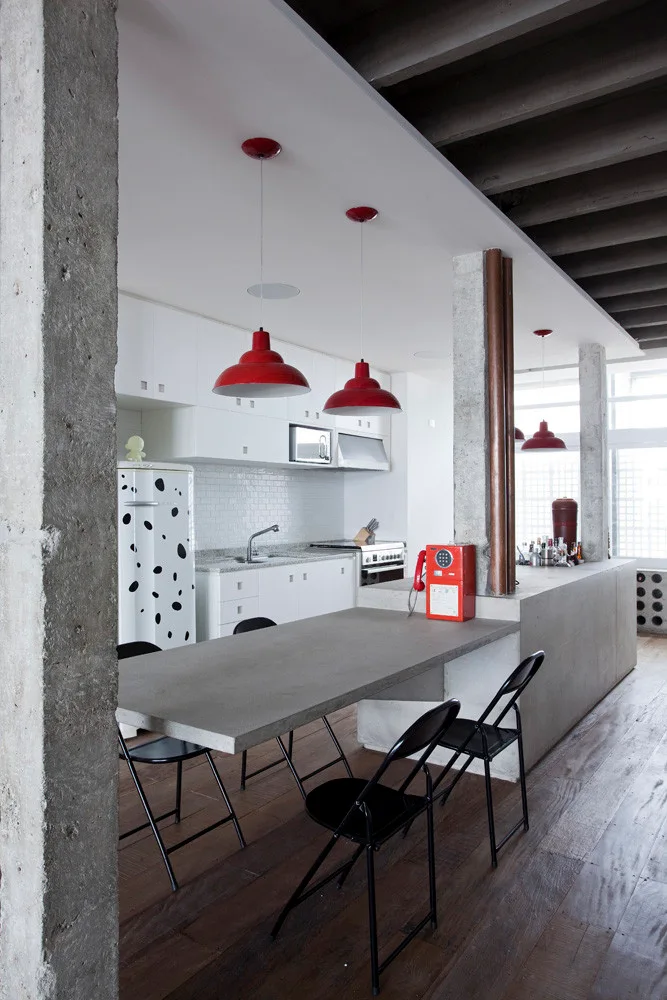 An industrial loft kitchen features a concrete breakfast bar with black folding chairs, white cabinetry, and bright red pendant lights.