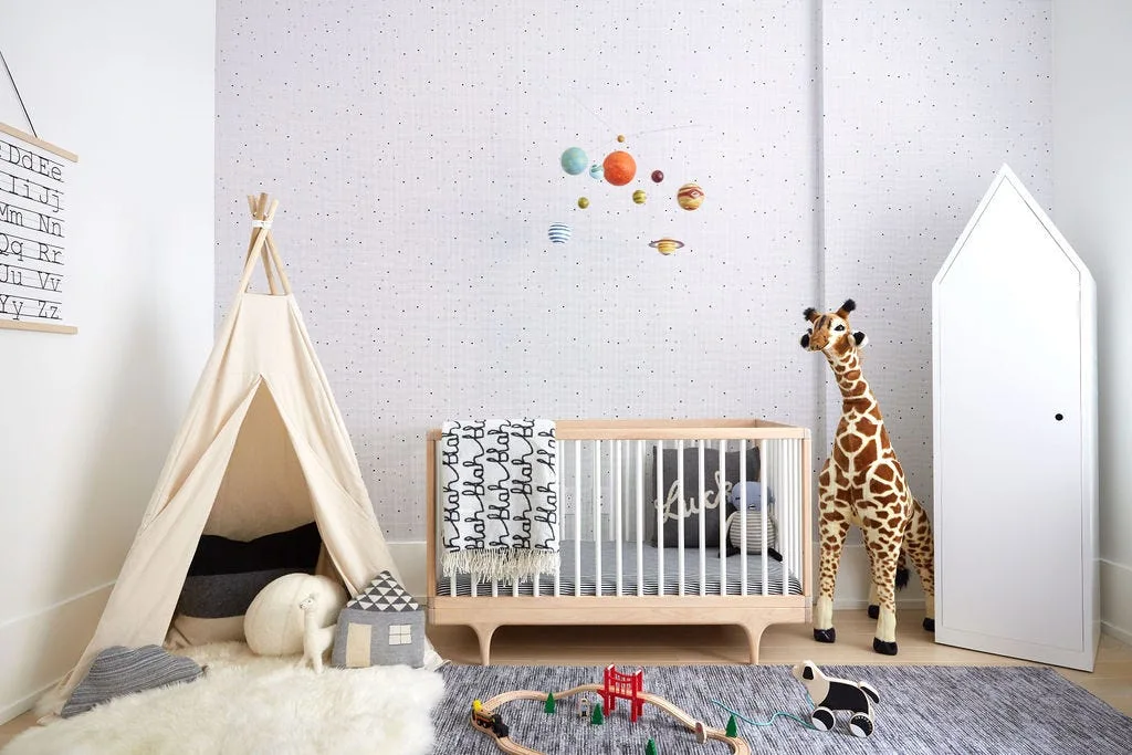 A Scandinavian-style nursery featuring a wooden crib, a canvas play teepee, a plush giraffe, and a tall house-shaped cabinet against dotted wallpaper.