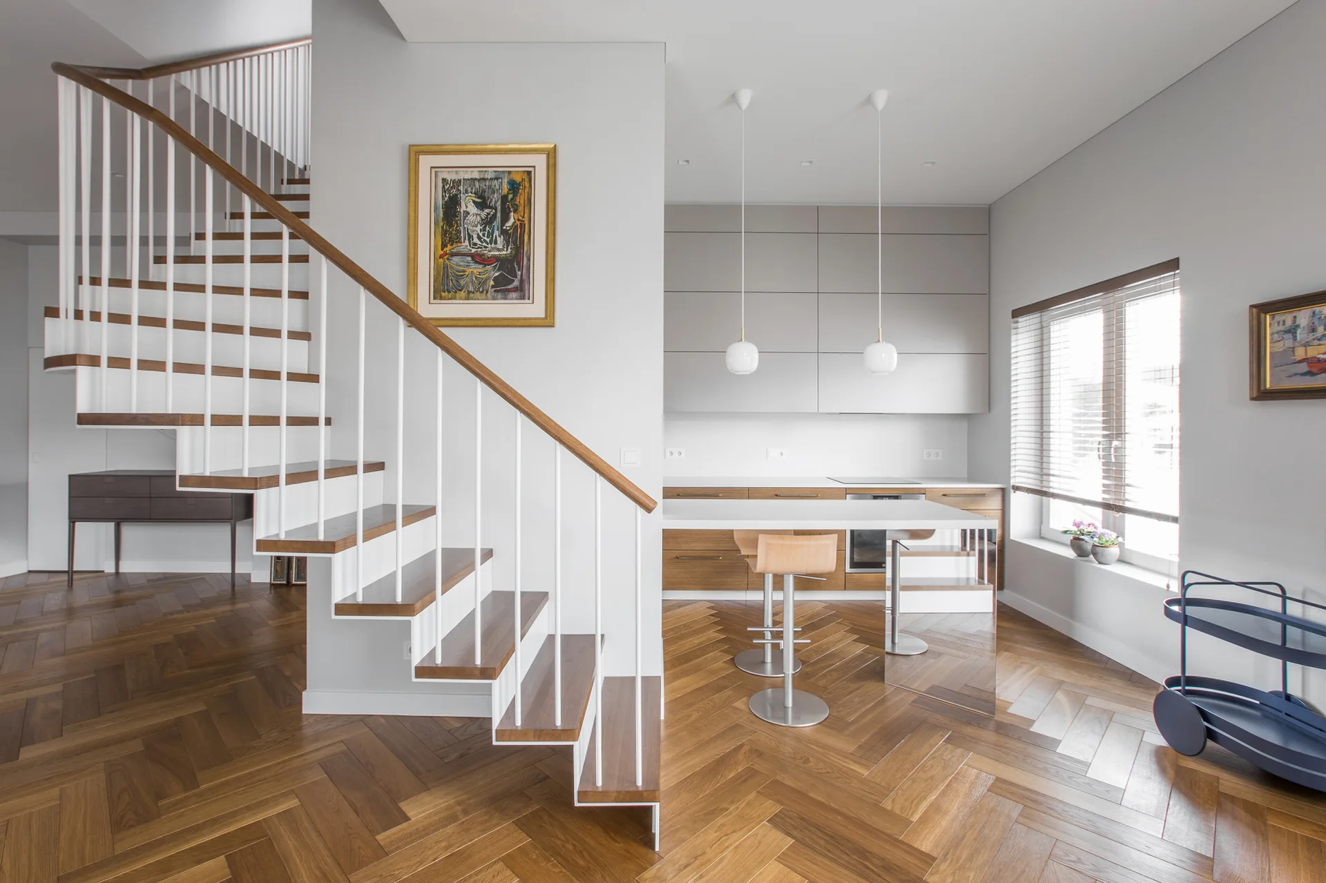 A modern open-plan kitchen features gray and wood cabinets, a white island, and a wooden staircase over herringbone pattern flooring.