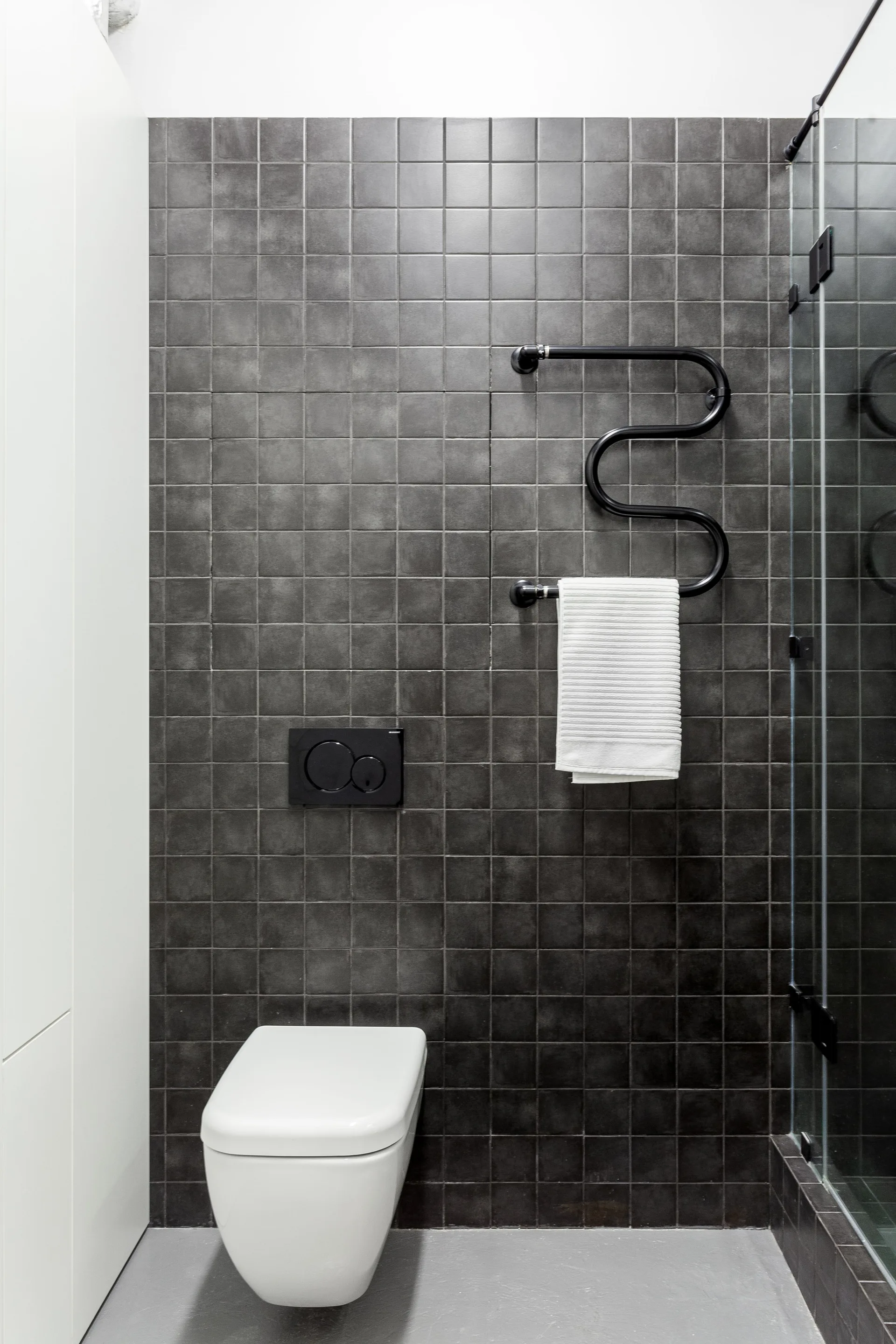 A modern bathroom featuring a white wall-mounted toilet against a dark grey square-tiled wall with a black curved towel warmer.
