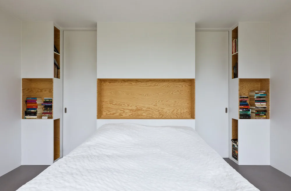 A bed centered in a white room against an integrated storage wall featuring a large recessed wood niche and matching side shelving for books.