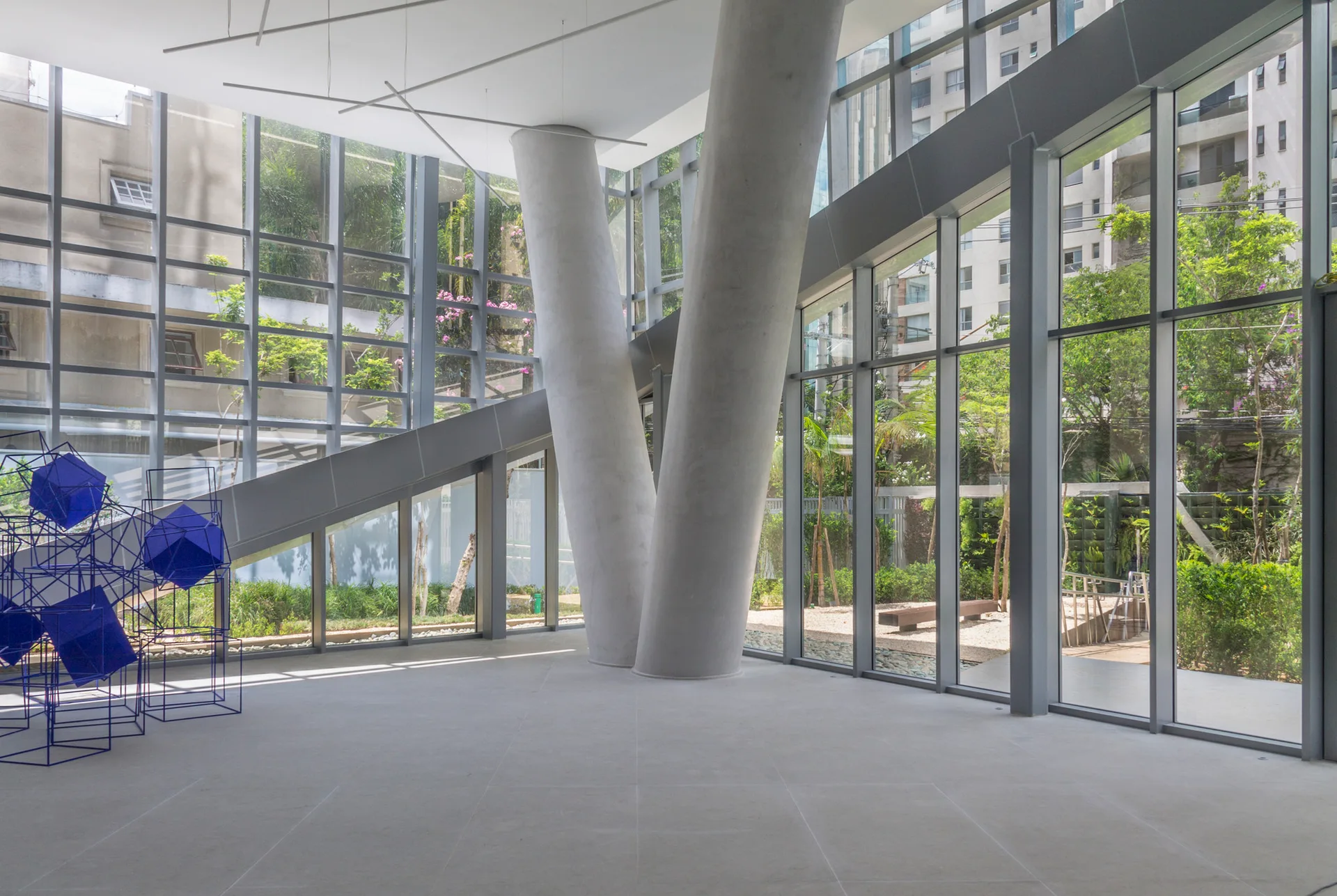 A minimalist glass-walled interior featuring two large angled concrete columns, a gray tiled floor, and a blue geometric wire sculpture.
