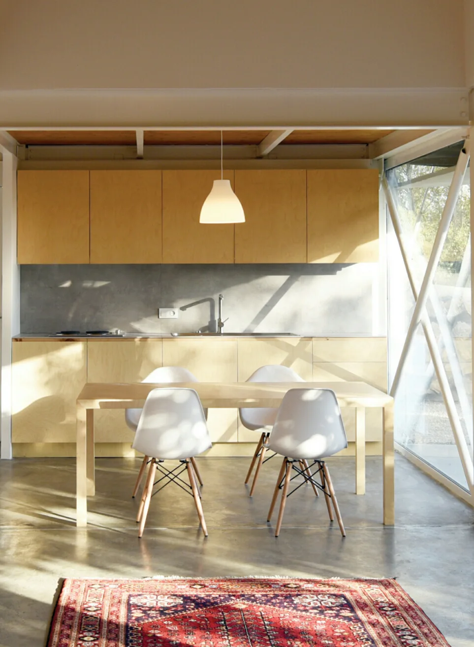 A minimalist kitchen features light plywood cabinets, a concrete floor, a light wood dining table with white chairs, and a traditional red patterned rug.