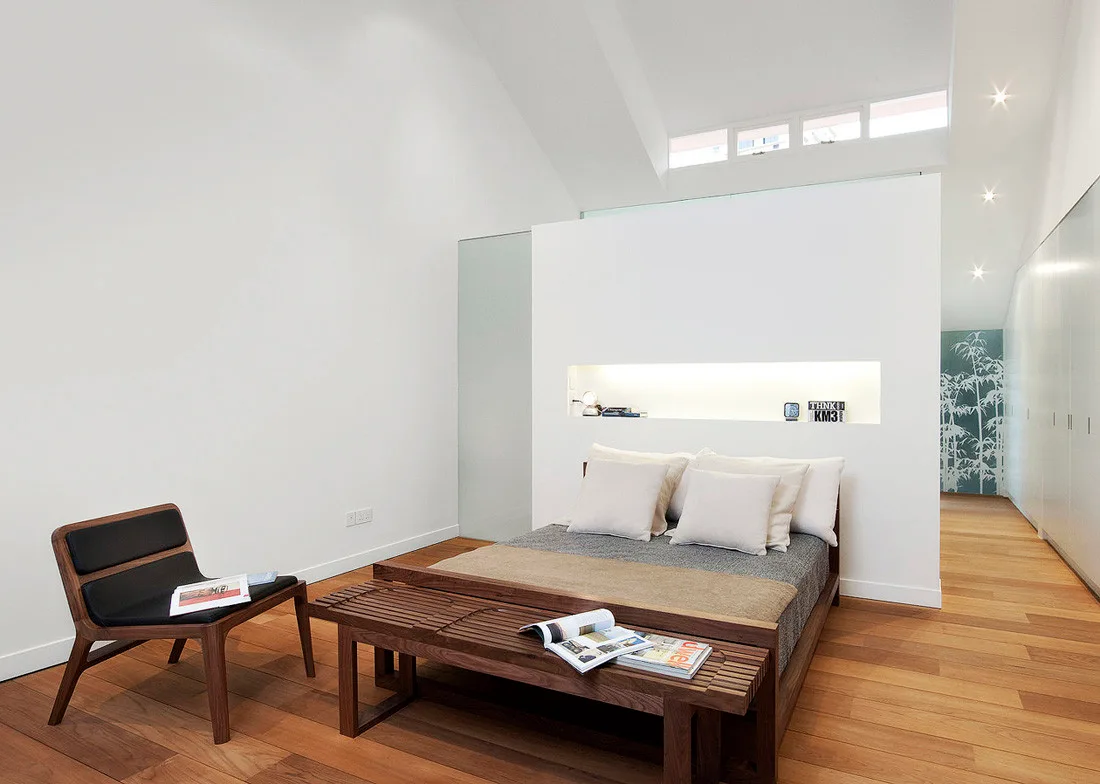 A minimalist modern bedroom featuring a low platform bed set against a white partition wall with an illuminated recessed niche and a wood bench.