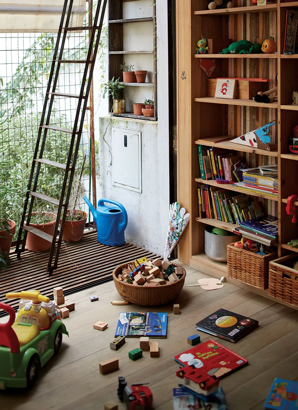 A kids playroom features a large wooden bookshelf next to a metal ladder leading to a sunlit balcony filled with potted plants.
