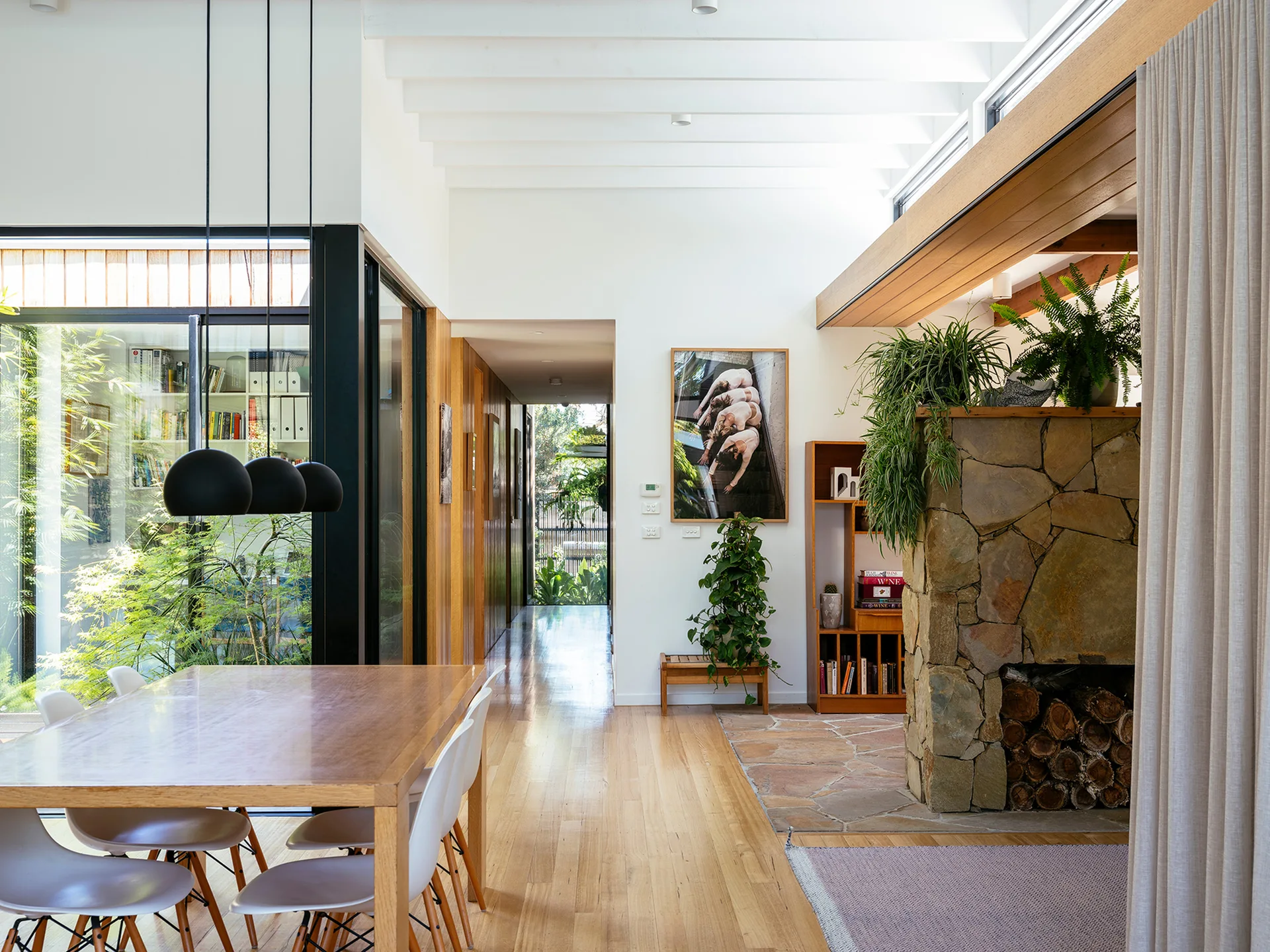 An open dining area features a wooden table with white chairs, a rugged stone fireplace, and white ceiling beams.