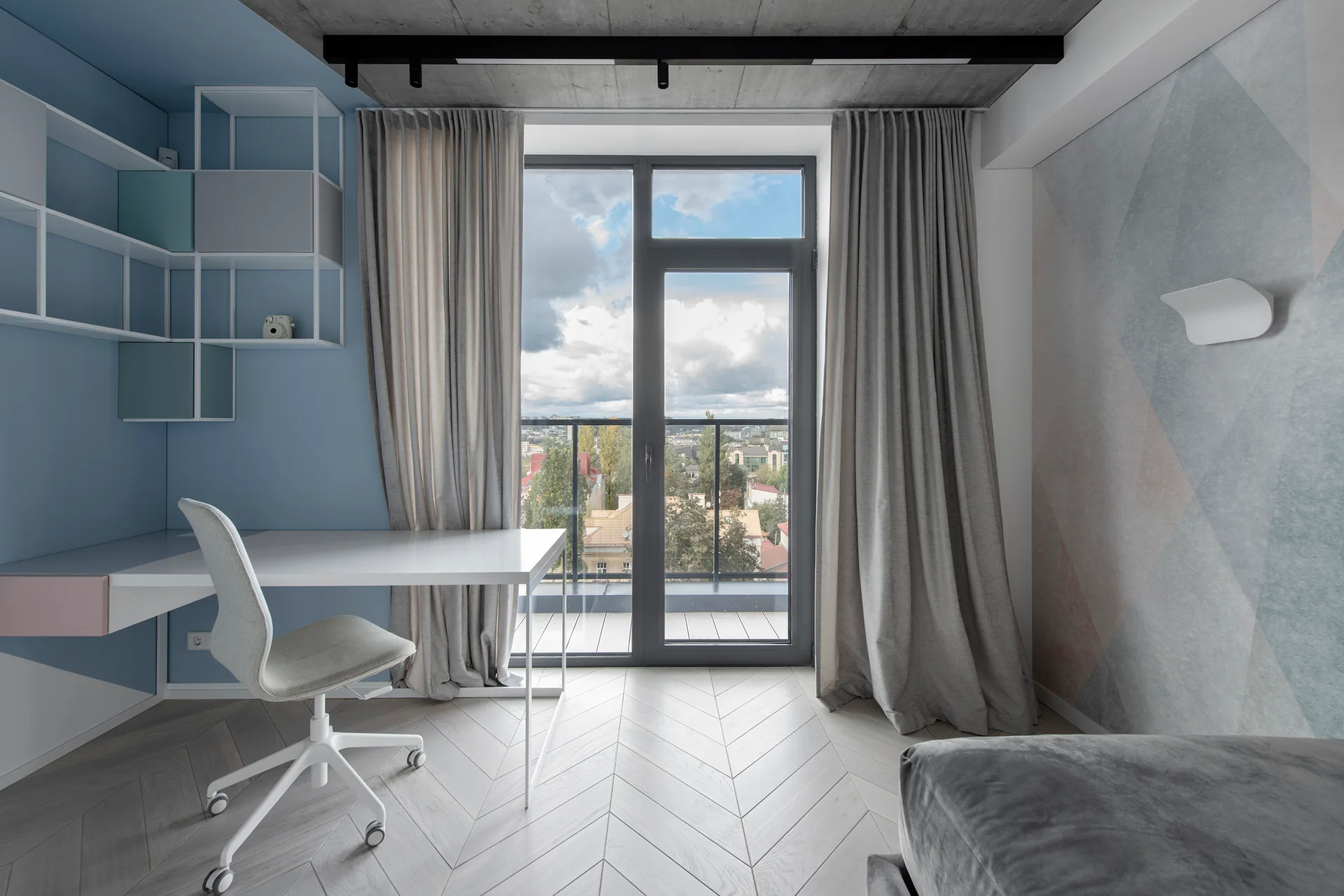 A modern home office features a white desk, grey chair, and blue wall with shelves, situated under an exposed concrete ceiling with a glass balcony door.