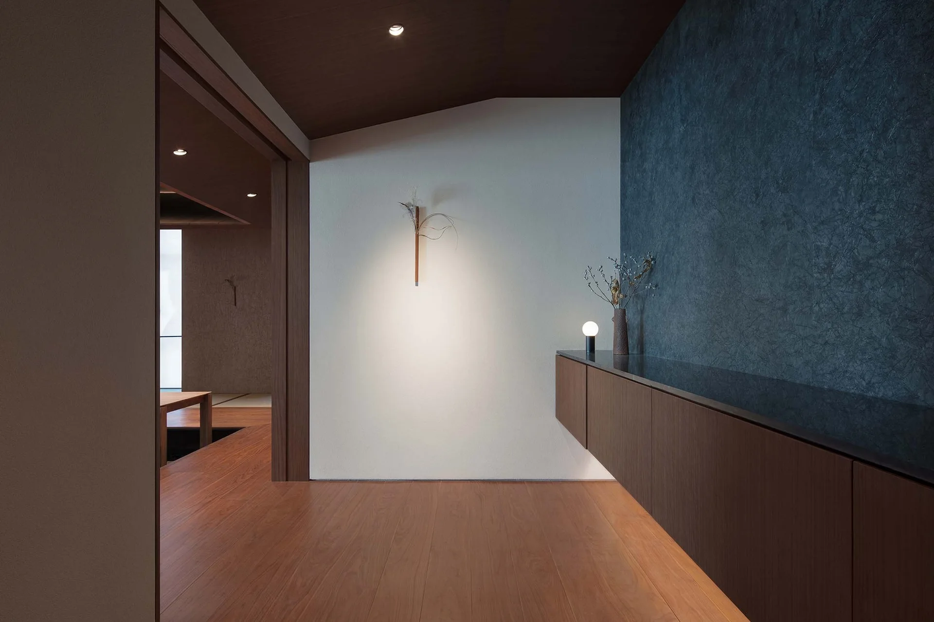 A minimalist hallway features wood plank flooring, a long floating wood console, a dark textured feature wall, and warm under-cabinet lighting.