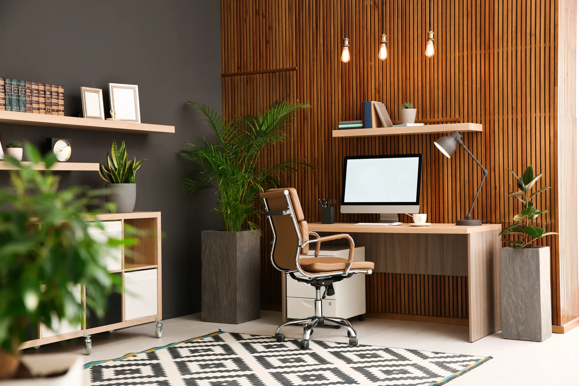 A modern home office features a wooden desk and brown leather chair against a vertical wood slat wall, complete with floating shelves and potted plants.