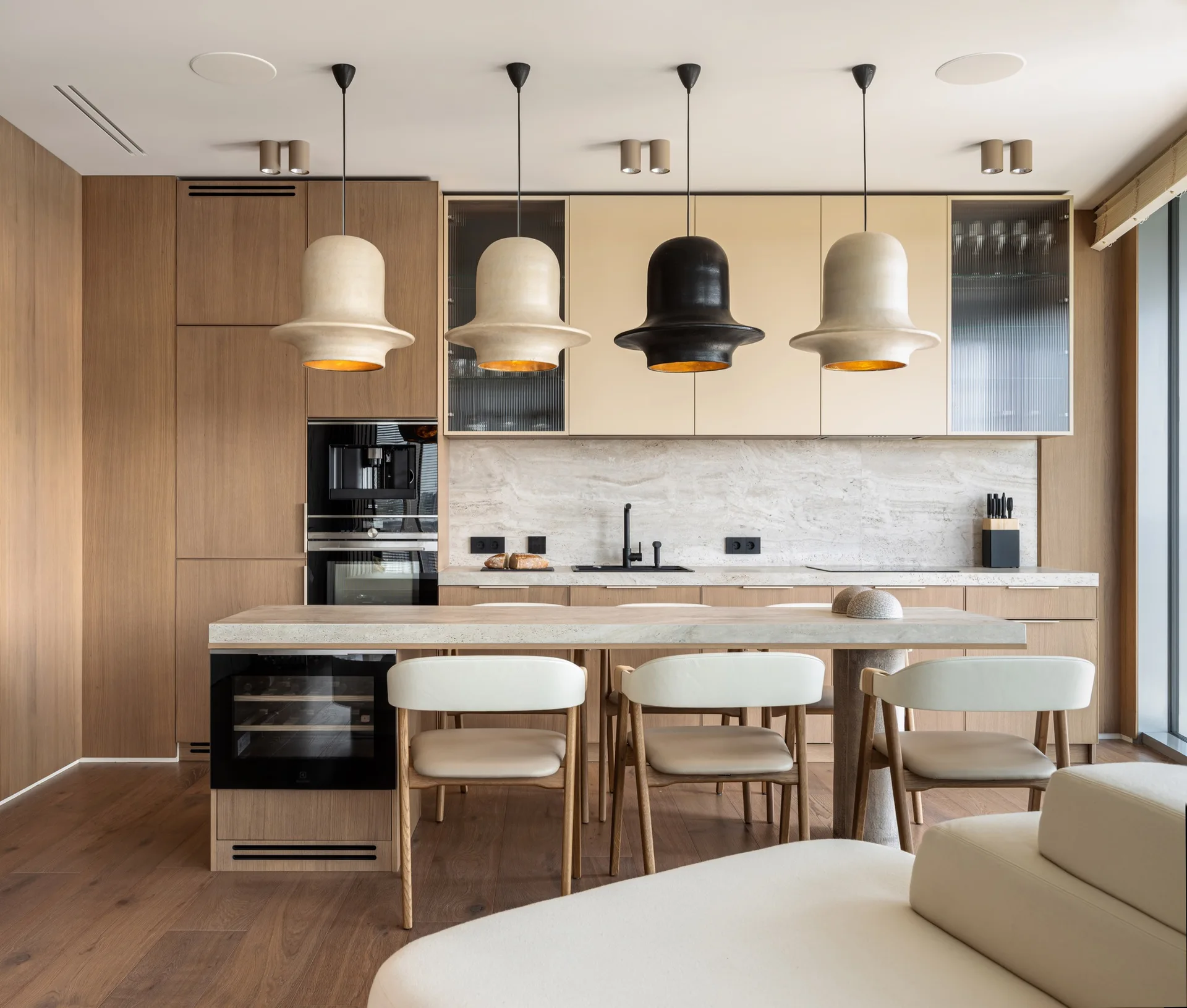 A modern kitchen features wood cabinetry, a stone-topped island with three white counter stools, and four large bell-shaped pendant lights hanging above.