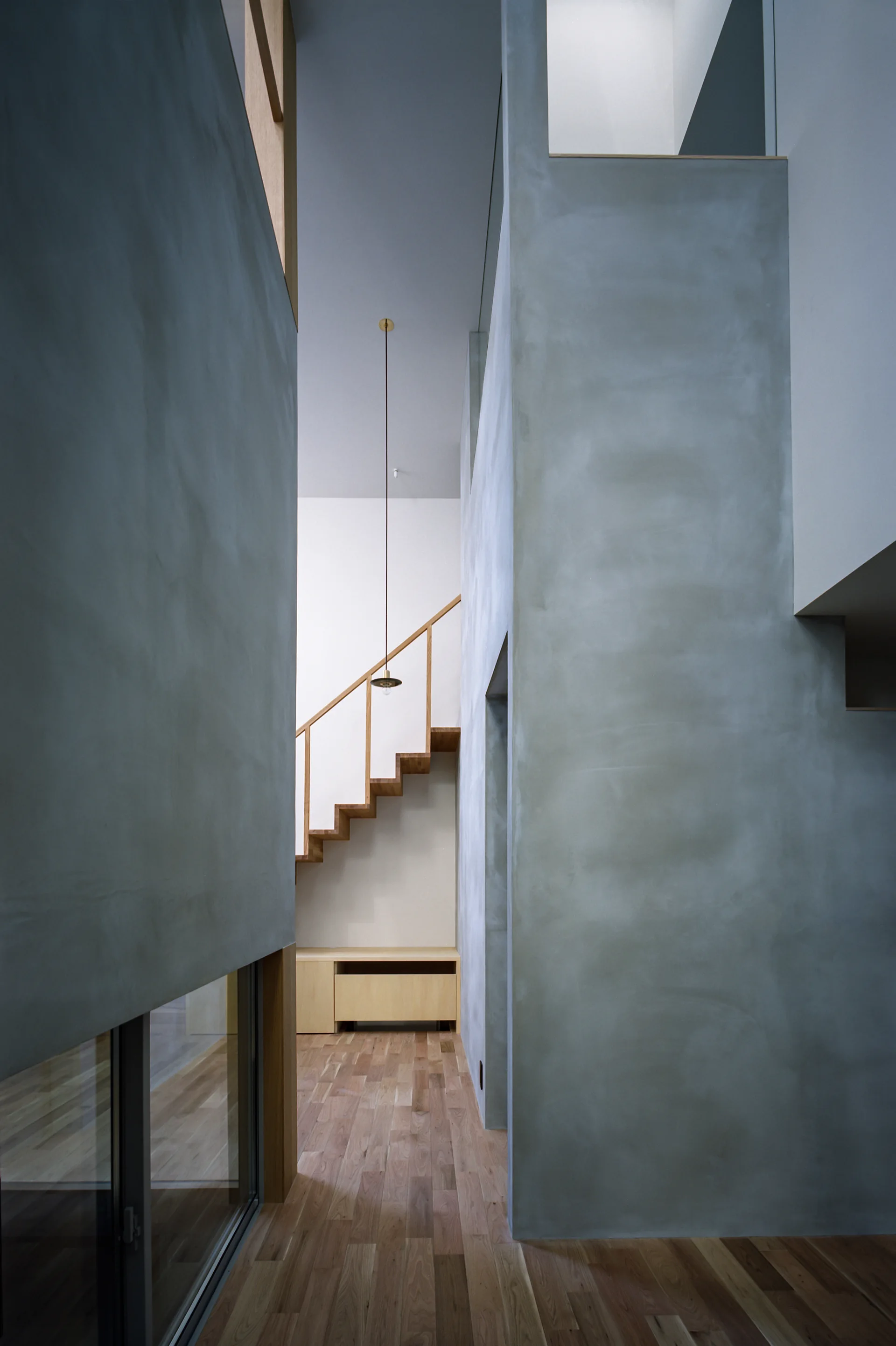 A minimalist hallway with tall gray plaster walls, a natural wood staircase with open risers, and a slender pendant light.