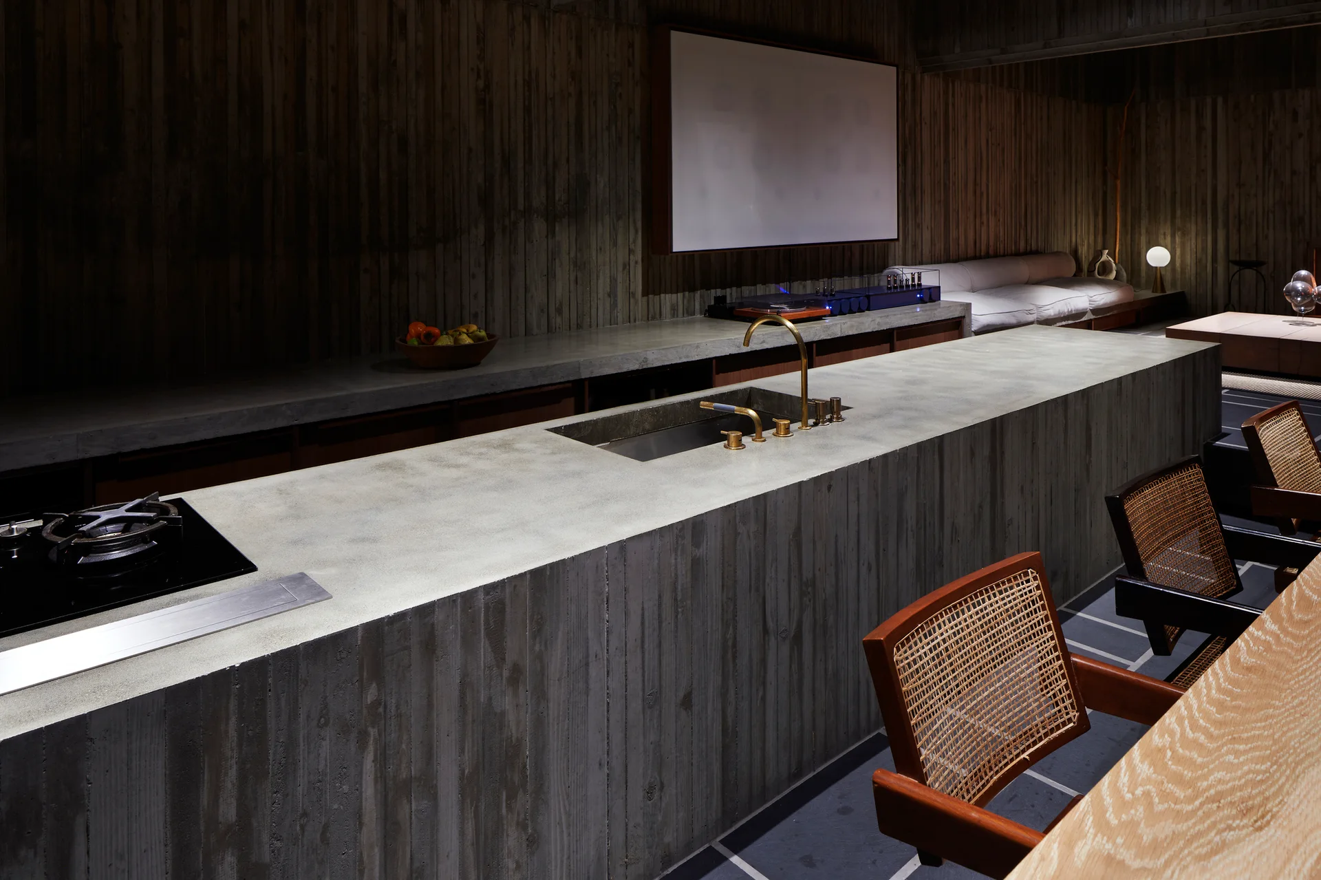 A minimalist kitchen features a massive board-formed concrete island with a gold faucet and a row of wooden cane-back chairs.