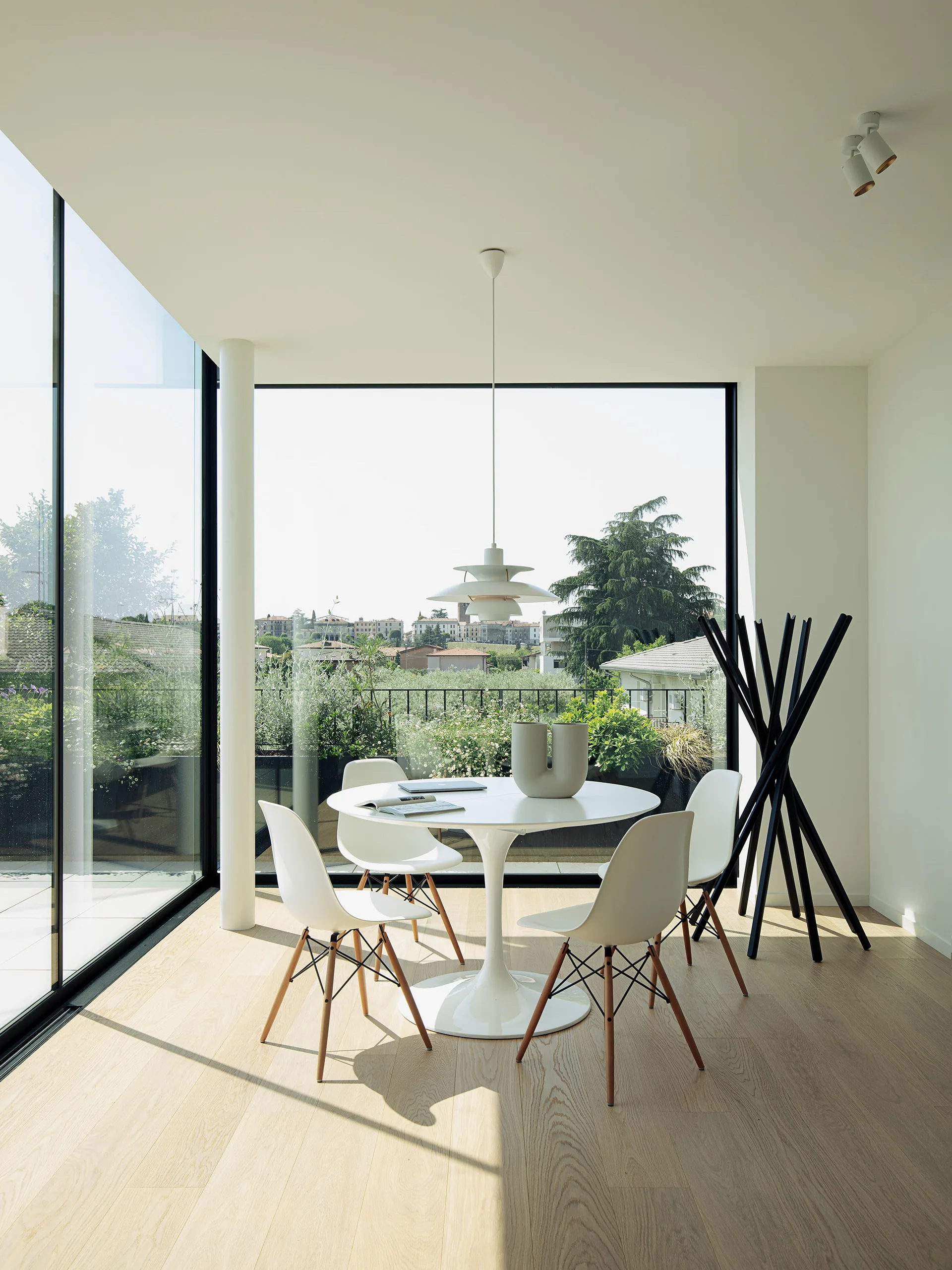 A white circular pedestal table and molded chairs sit on light wood floors in a dining room next to large floor-to-ceiling windows.