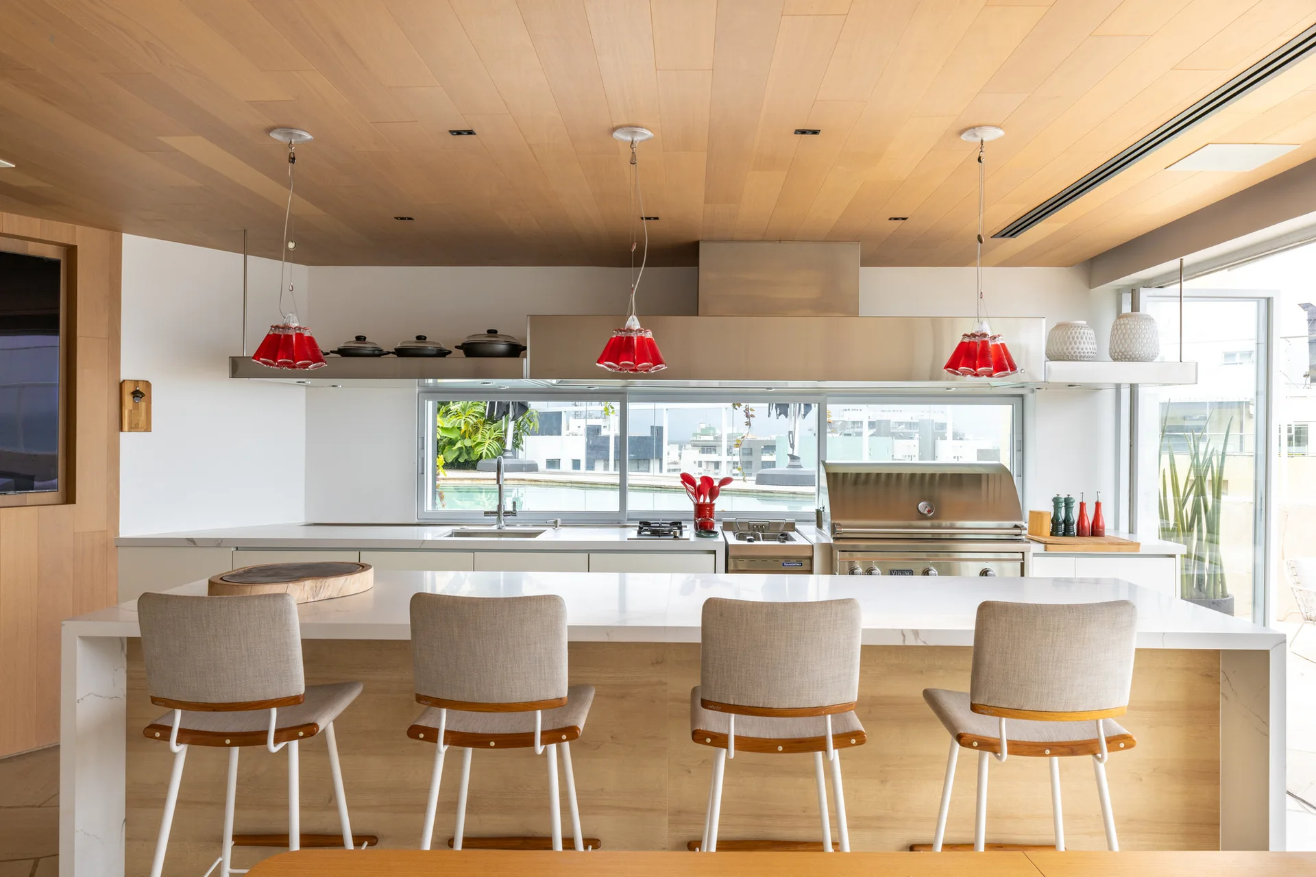 A modern kitchen features a marble-topped island with four bar stools, three red glass pendant lights, and a wood plank ceiling.