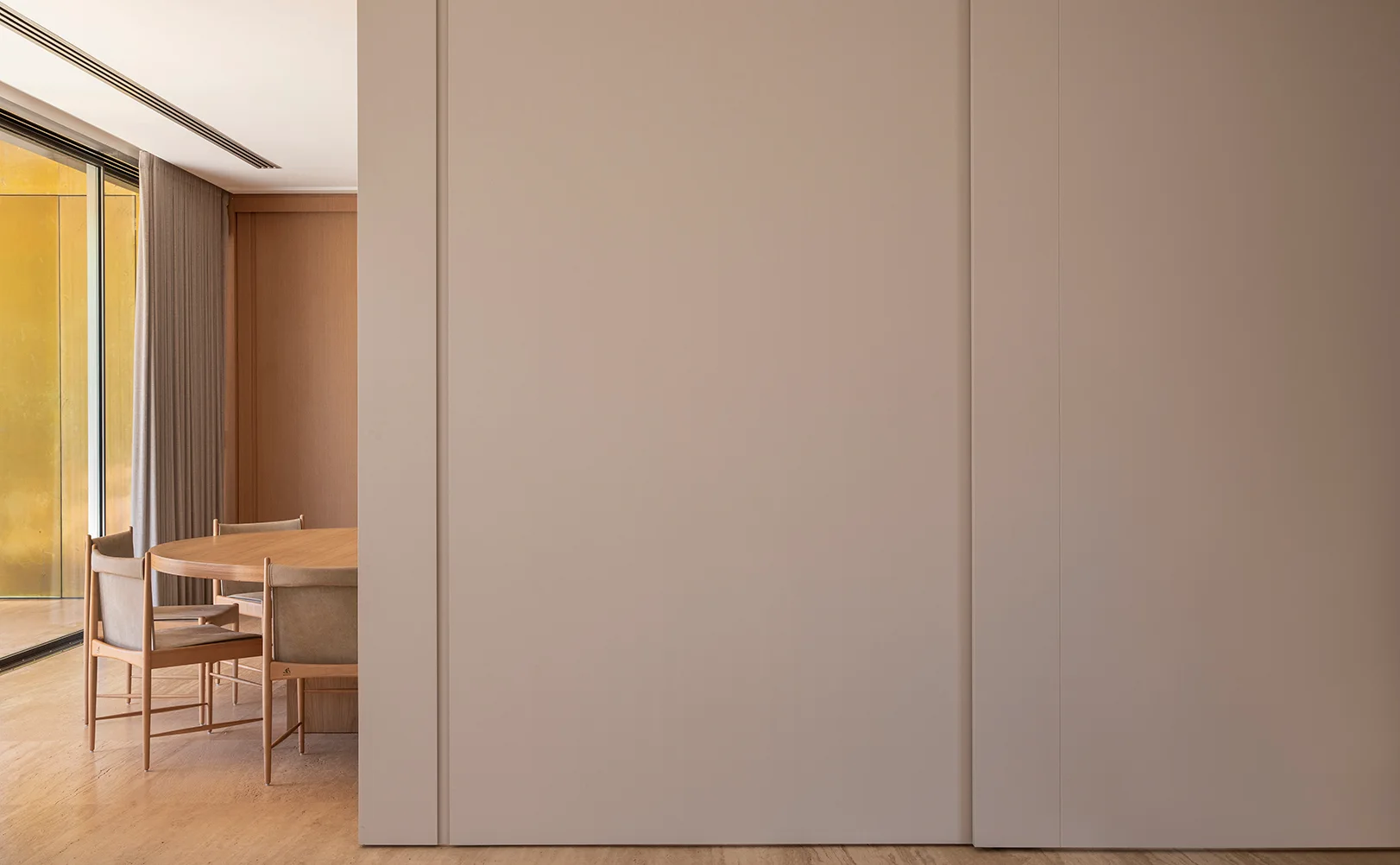 A minimalist dining area with a round wooden table and chairs is partially visible behind a large, flat beige wall panel next to sliding glass doors.