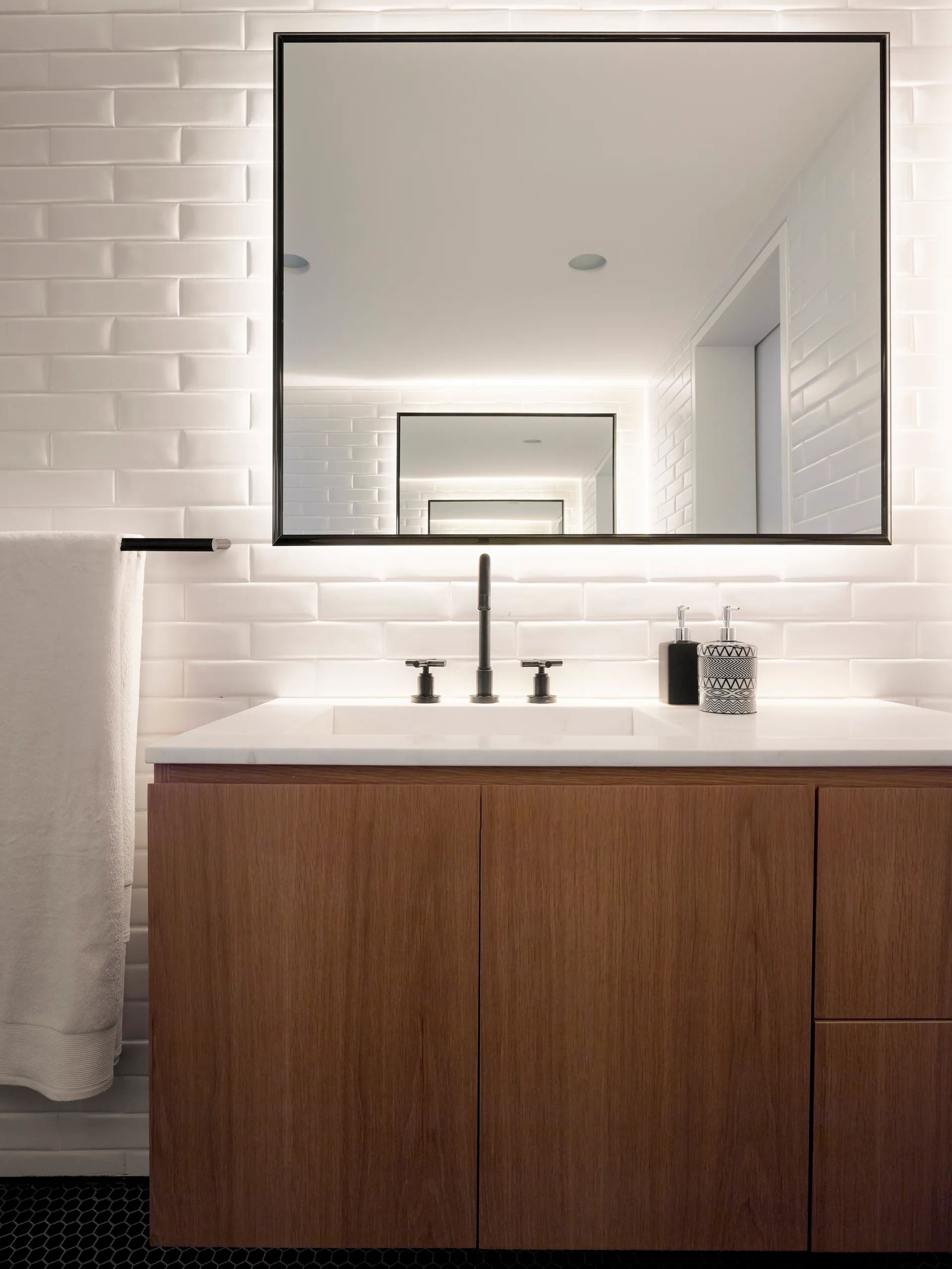 A modern wood bathroom vanity features a white countertop, matte black faucet, and a large backlit mirror against a white subway tile wall.