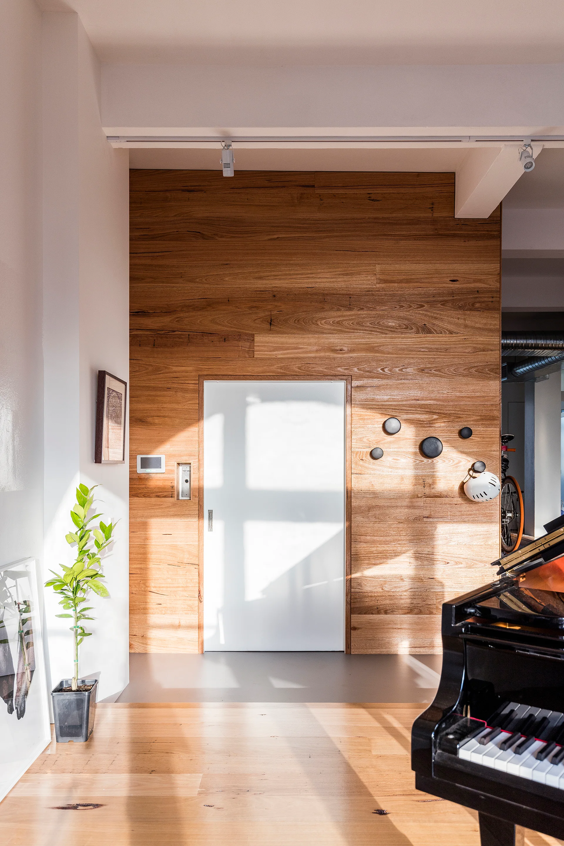 An entryway features a warm horizontal wood paneled wall with a white door, a partial view of a black grand piano, and light hardwood floors.