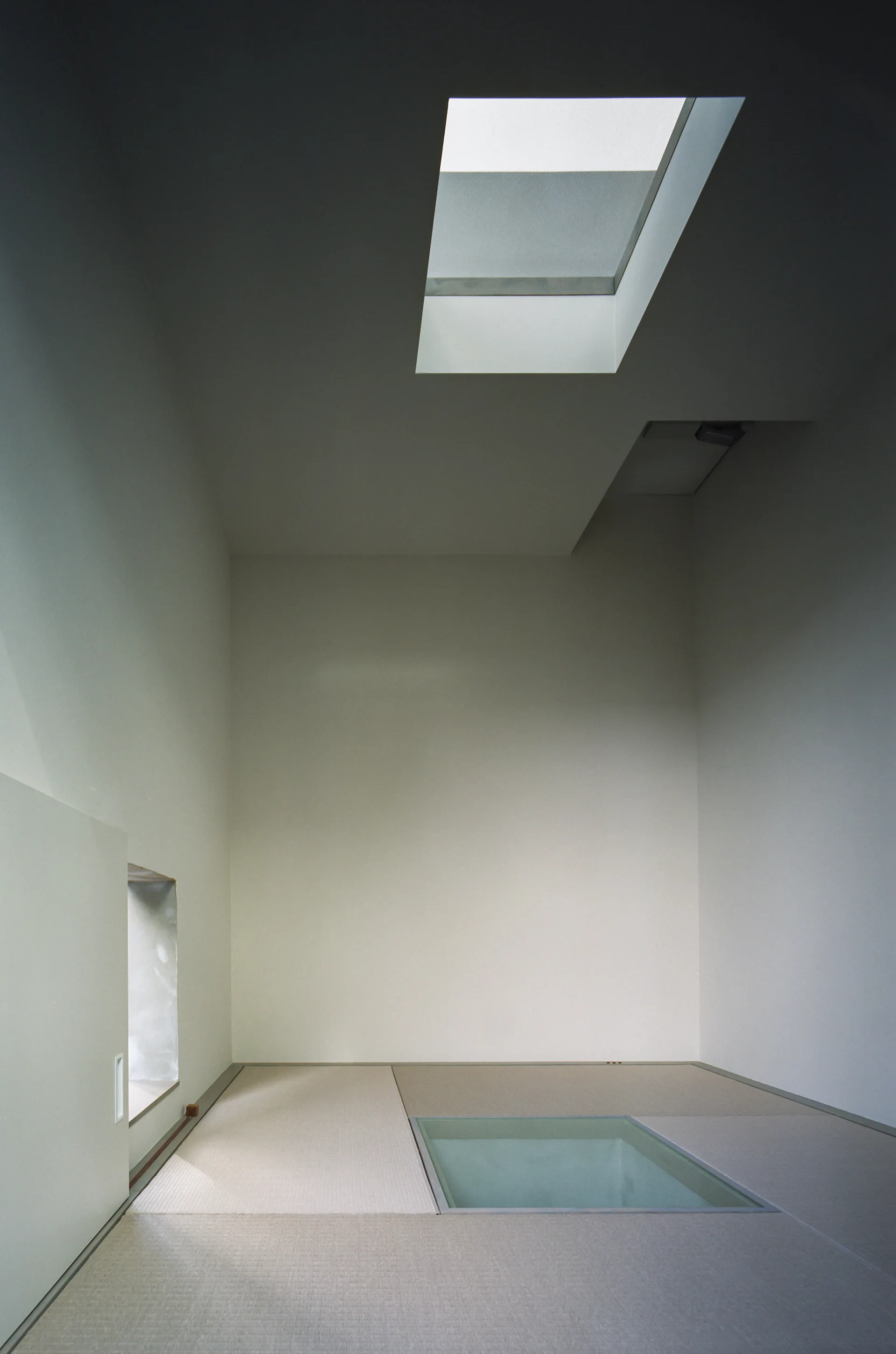 A minimalist room with white walls, textured woven floor mats, a ceiling skylight, and a glass floor panel.