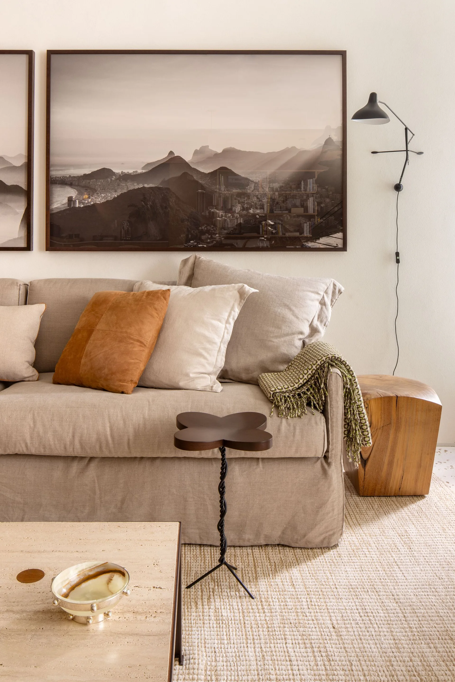 A beige slipcovered linen sofa with accent pillows sits near a travertine coffee table, a sculptural metal side table, and a large framed landscape photo.