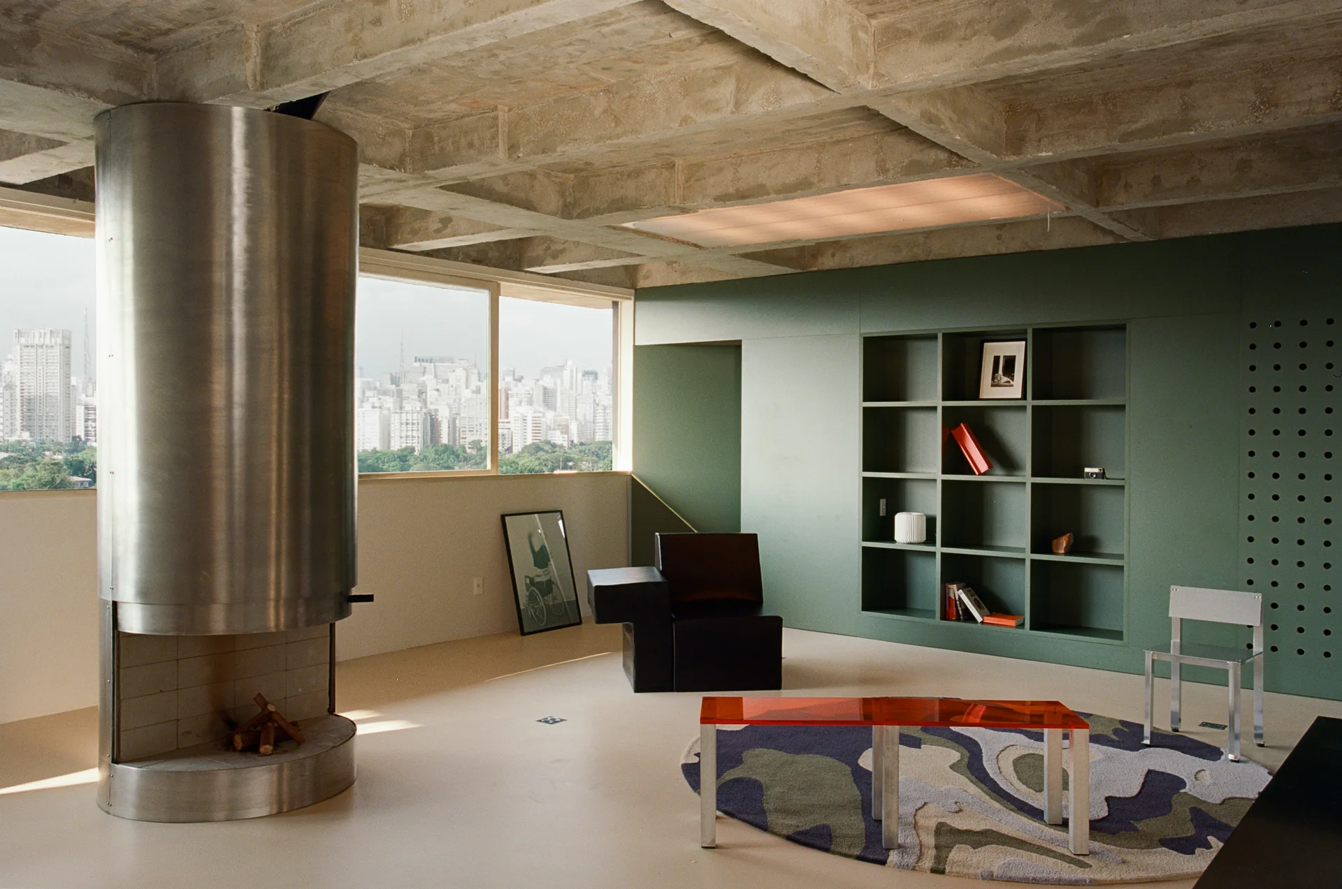 A minimalist living room with an exposed concrete waffle ceiling, a cylindrical stainless steel fireplace, and an olive green built-in wall shelf.