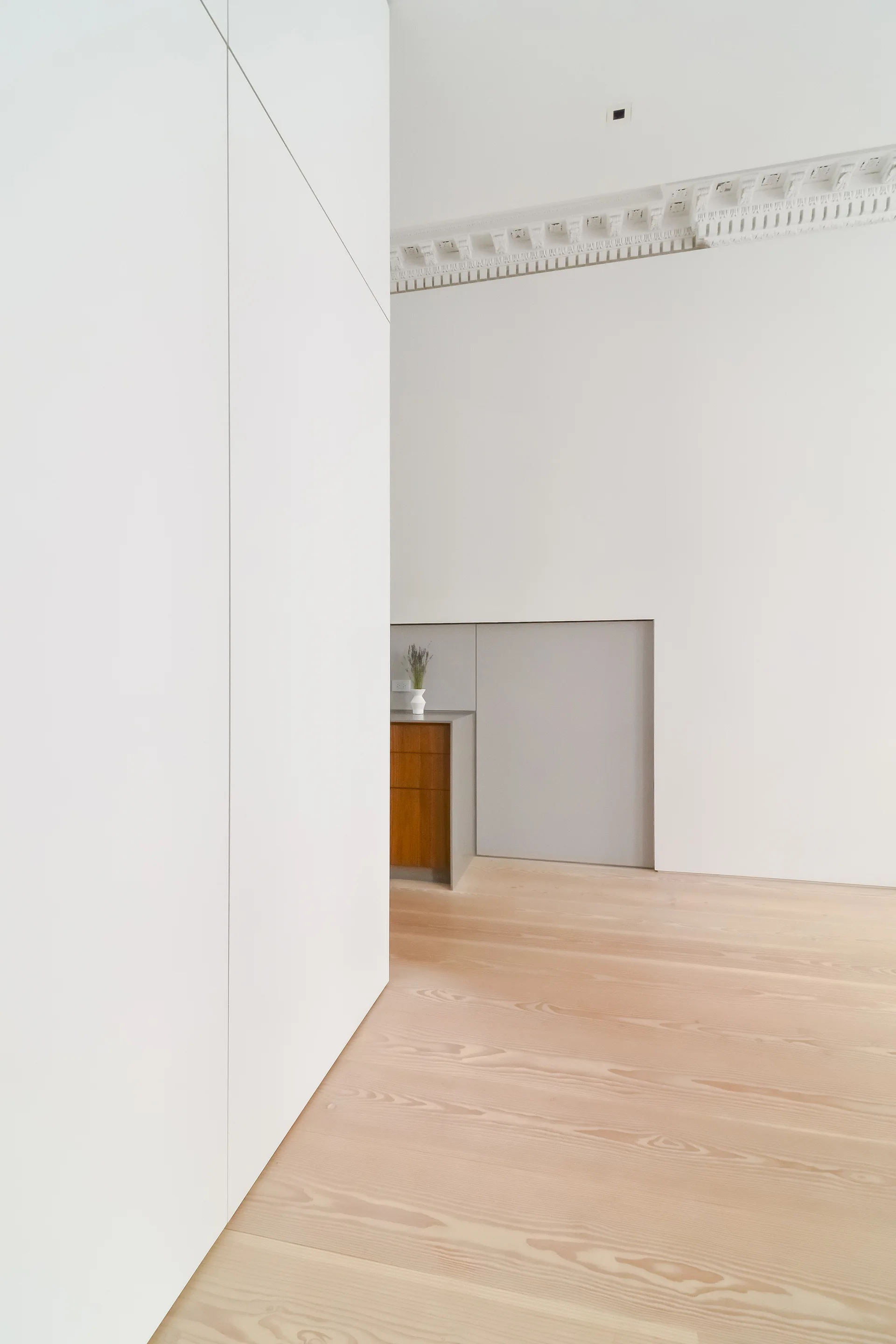 A minimalist interior space features a flush white storage wall, light wood plank floors, and ornate classical crown molding.