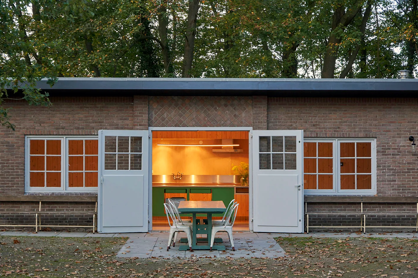 An outdoor dining table with white metal chairs sits on a stone patio in front of a brick building with open doors showing a lit kitchen.