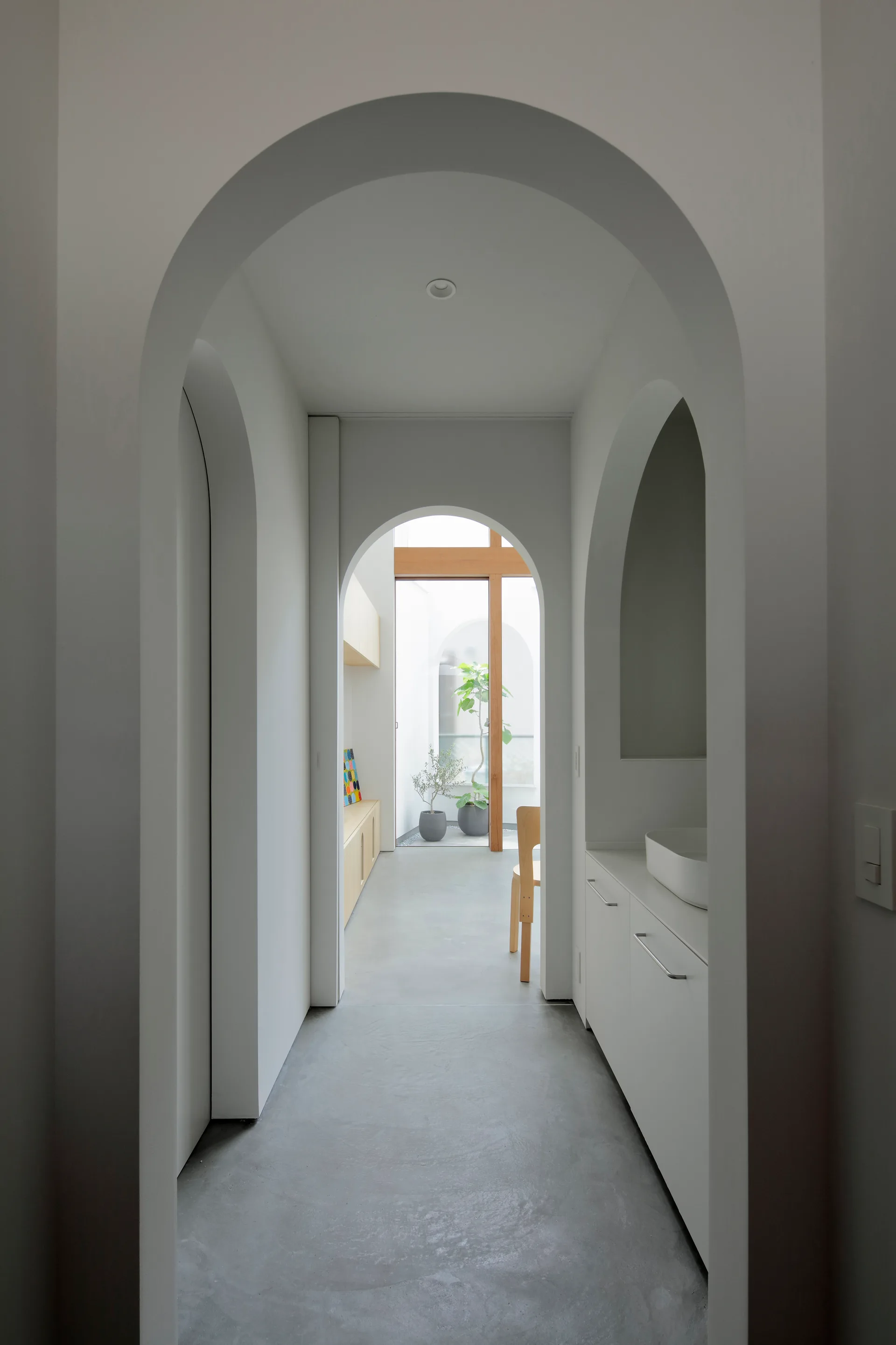 A minimalist hallway features nested white plaster archways, smooth grey concrete flooring, a built-in vanity, and a view of a bright courtyard.