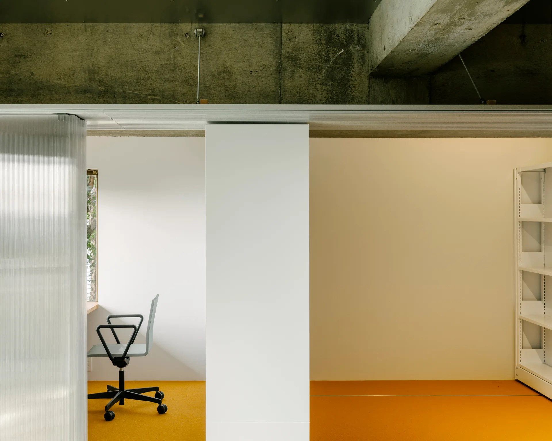 A minimalist home office features bright orange flooring, a translucent fluted partition screen, a grey desk chair, and an exposed raw concrete ceiling.