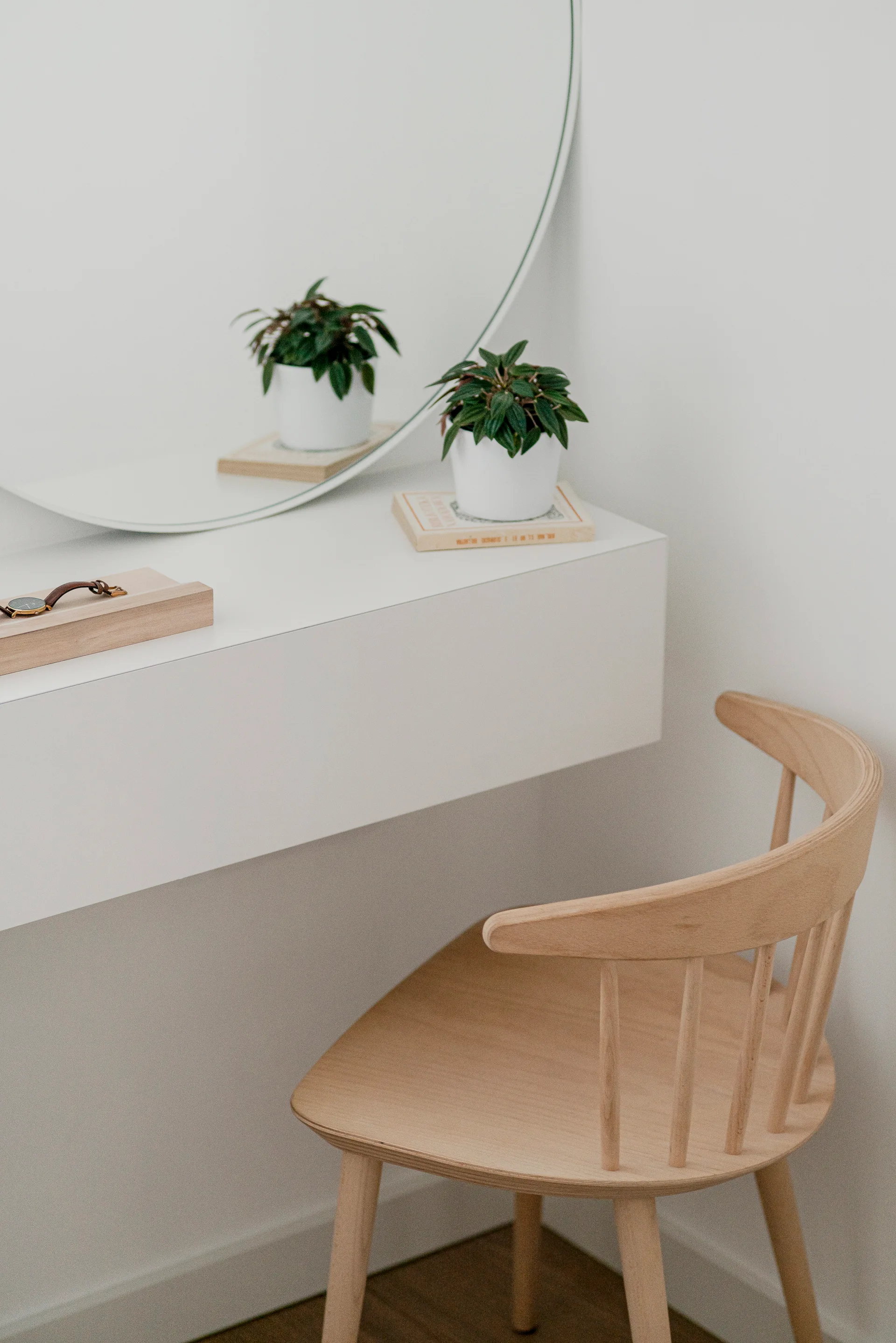 A minimalist white floating vanity features a round mirror, a small potted plant, and a light wood spindle-back chair.