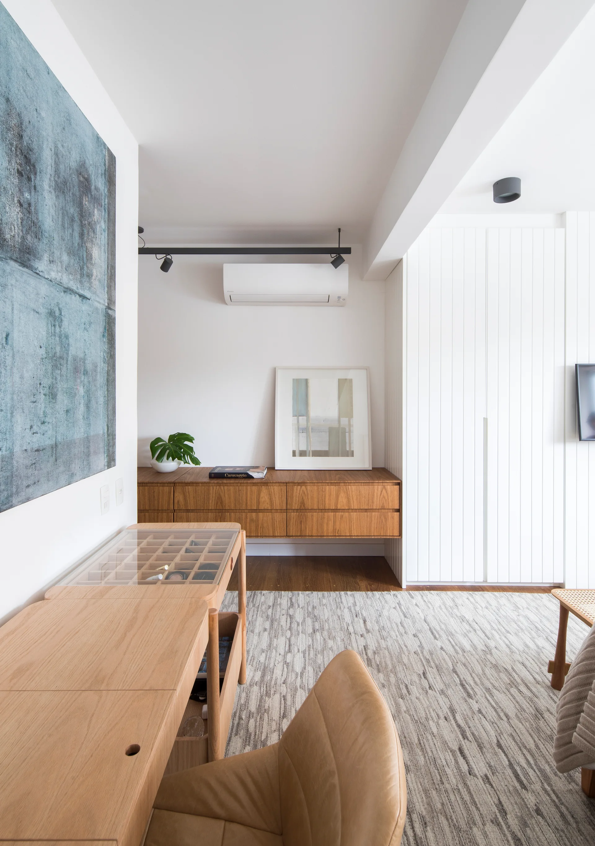 A modern bedroom featuring a light wood desk, a tan leather chair, a floating wooden credenza, and vertical white built-in cabinets.