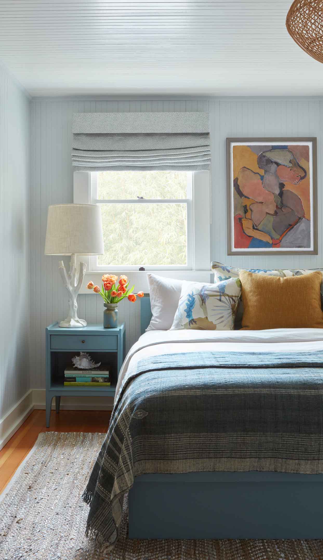 A coastal bedroom features light blue beadboard walls, a matching blue bed and nightstand, a white branch-style table lamp, and a textured woven rug.